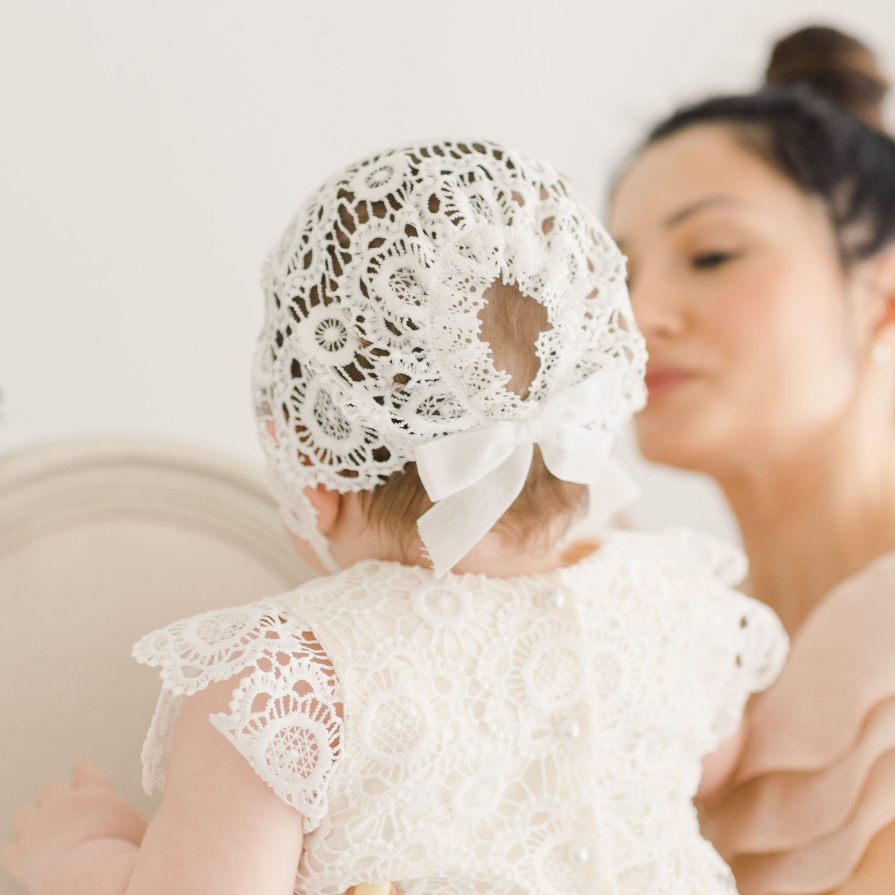 Close-up of the intricate floral lace bonnet and gown of the Poppy Christening Gown, showing delicate details and a ribbon tie at the back, with a blurred figure in the background.