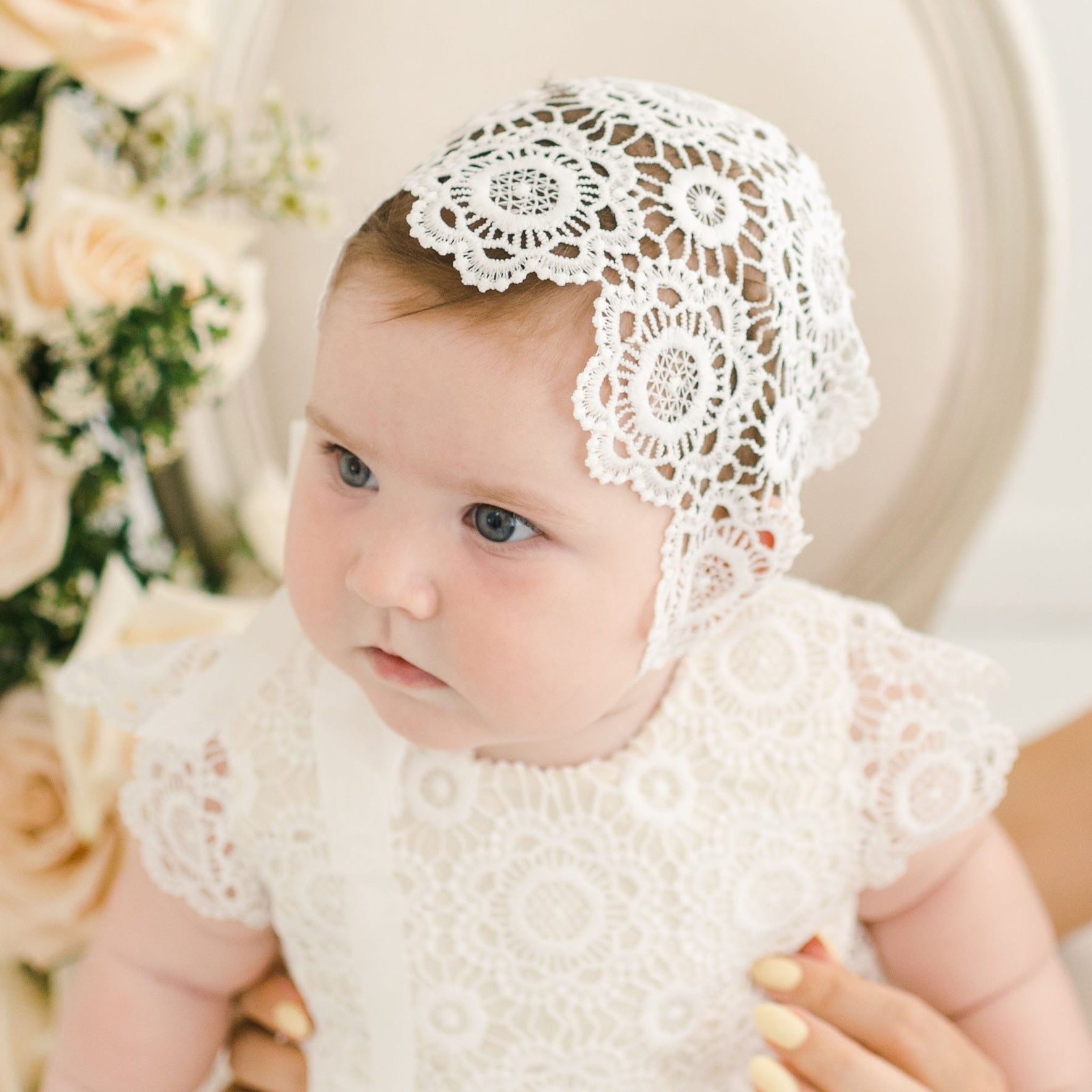 Close-up of a baby in the Poppy Christening Gown and bonnet, featuring detailed floral lace patterns and ivory accents, framed by a neutral chair and soft peach floral arrangements.