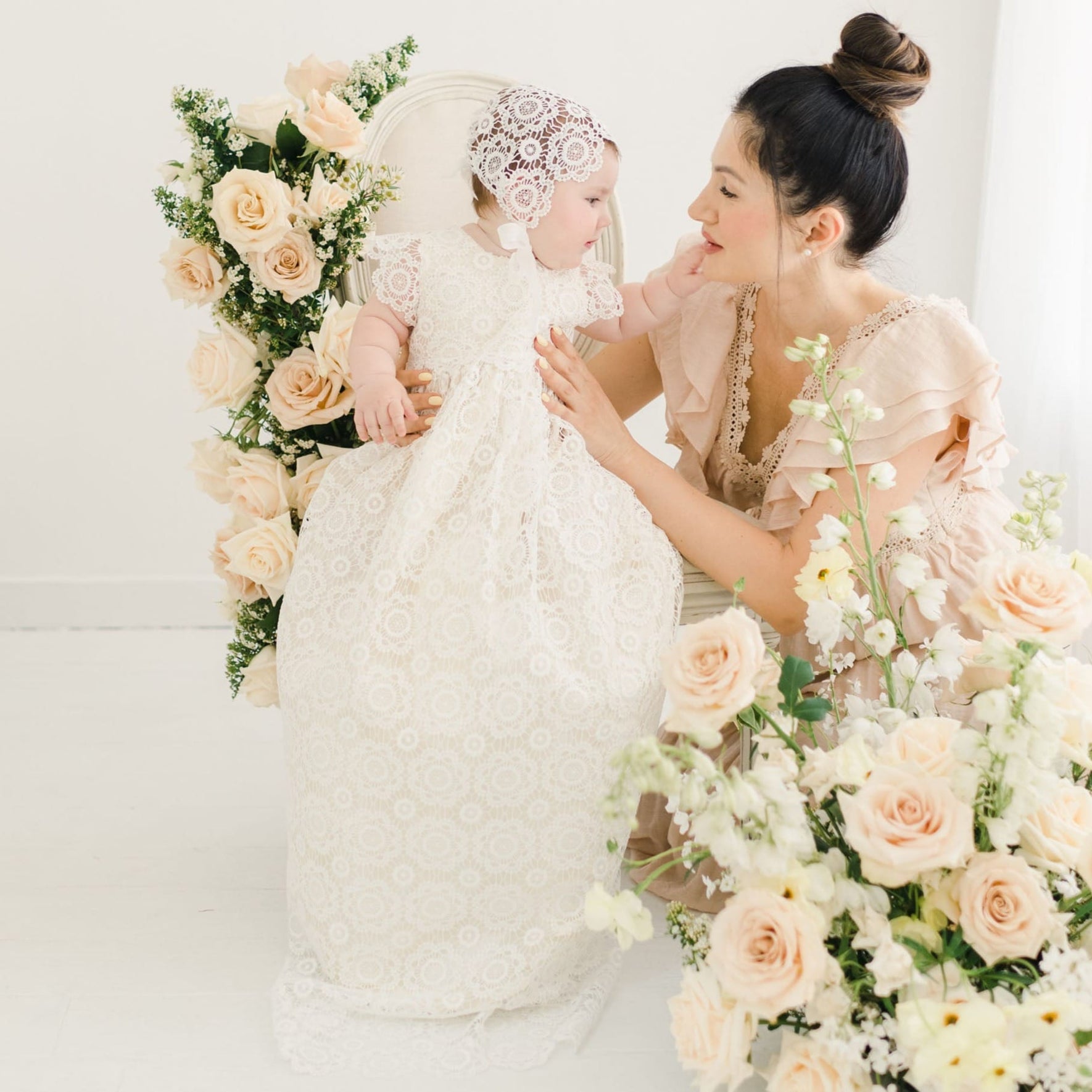 Baby girl dressed in the Poppy Christening Gown and Bonnet, seated on a chair surrounded by soft peach and cream floral bouquets, with her mother kneeling beside her in a gently lit setting.