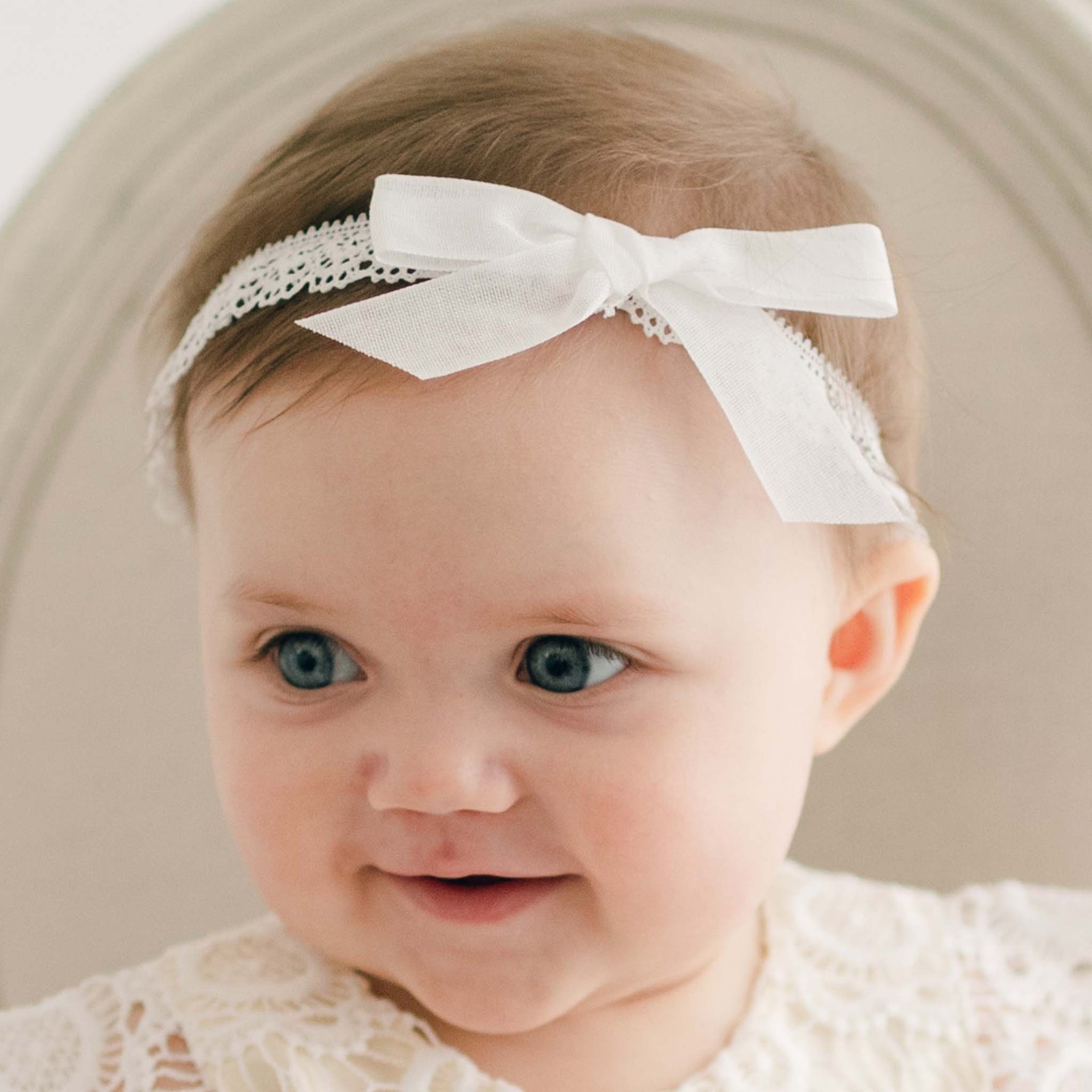 Side view of the Poppy Christening Headband, featuring a white bow and delicate lace trim, worn by a baby dressed in a lace gown.