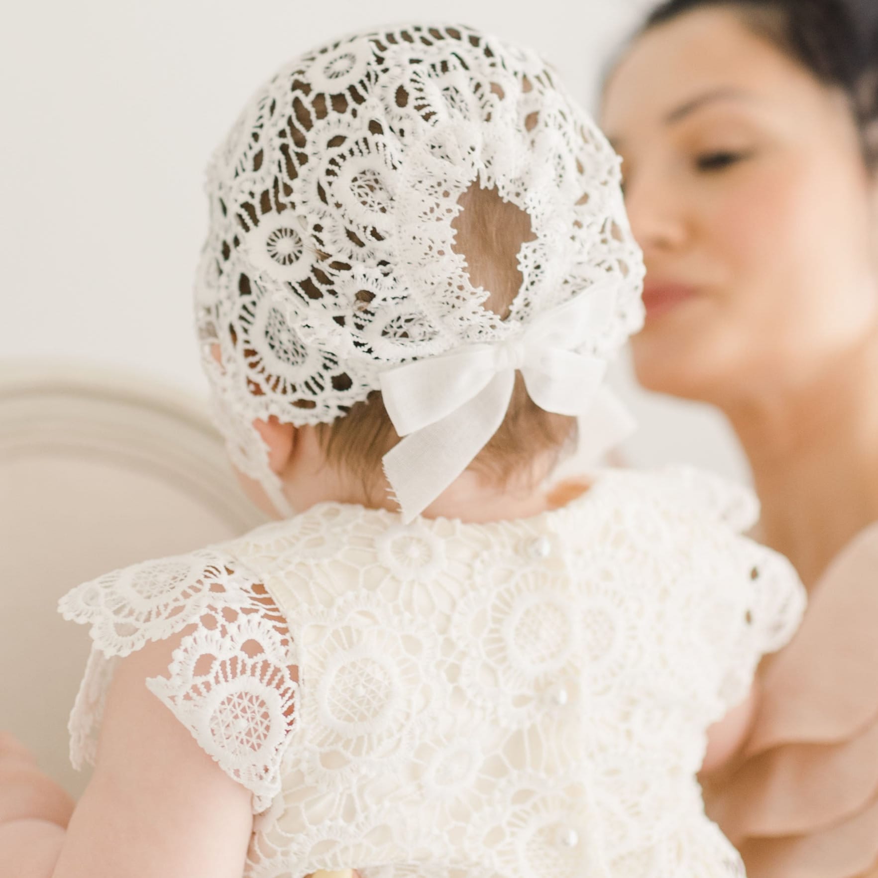 Rear view of the Poppy Blessing Dress and matching lace bonnet, showcasing delicate lacework and a satin bow as the baby is held by her mother.