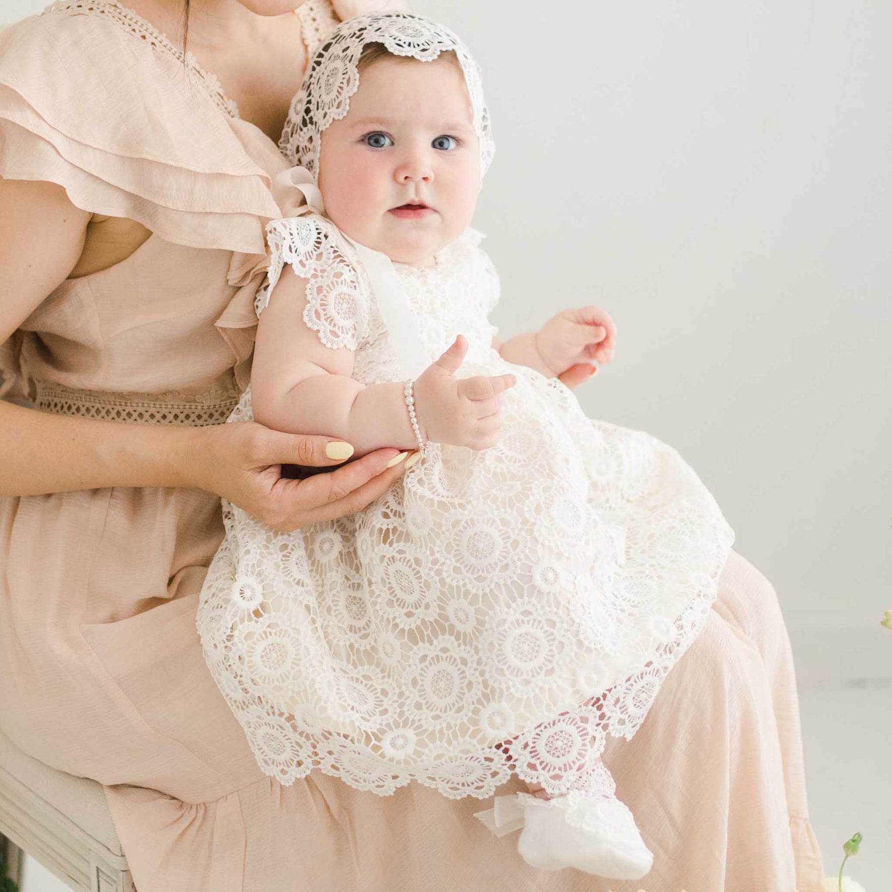 Baby girl dressed in the Poppy Christening Gown with lace detail and matching bonnet, held by her mother. The delicate floral lace and ribbon accents are featured.