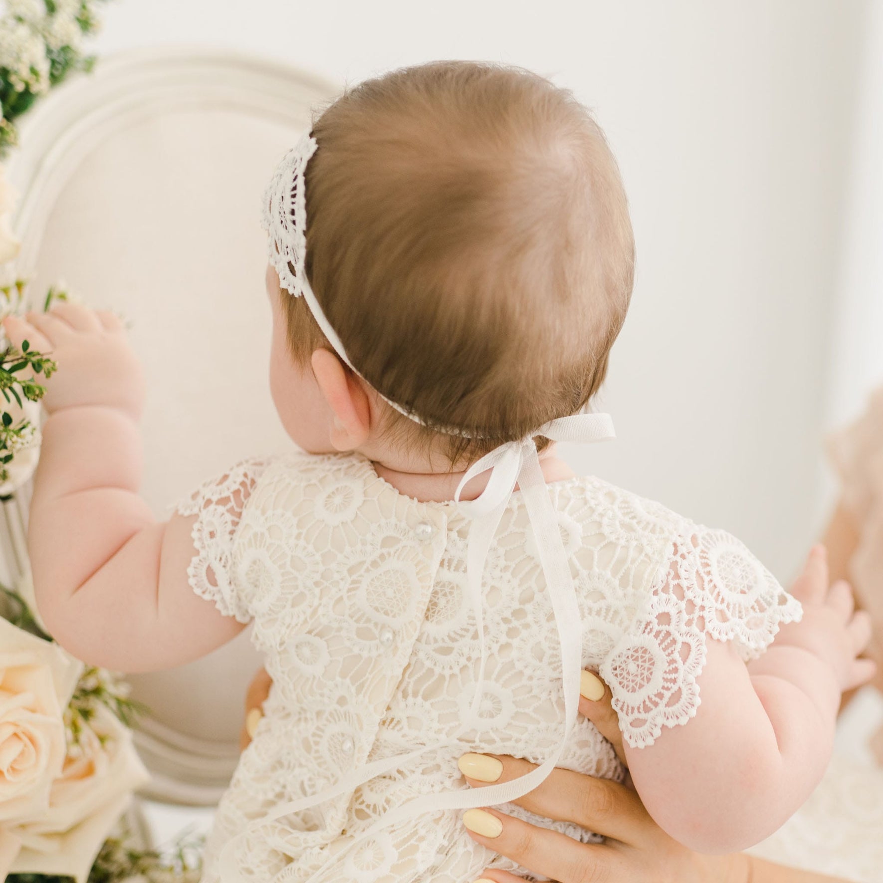 Back view of a baby wearing the Poppy Lace Headband, tied with a delicate white ribbon, and the coordinating lace gown with intricate floral patterns and pearl buttons.