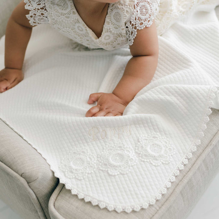 A close-up of a baby wearing an ivory lace baby dress while lying on an ivory textured blanket draped over a light beige upholstered chair.