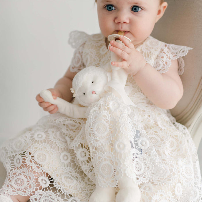 An infant in the Poppy Blessing Dress holding the Poppy Silly Lamb Buddy Pacifier Holder, featuring a plush lamb adorned with delicate lace matching the gown, holding a pacifier.