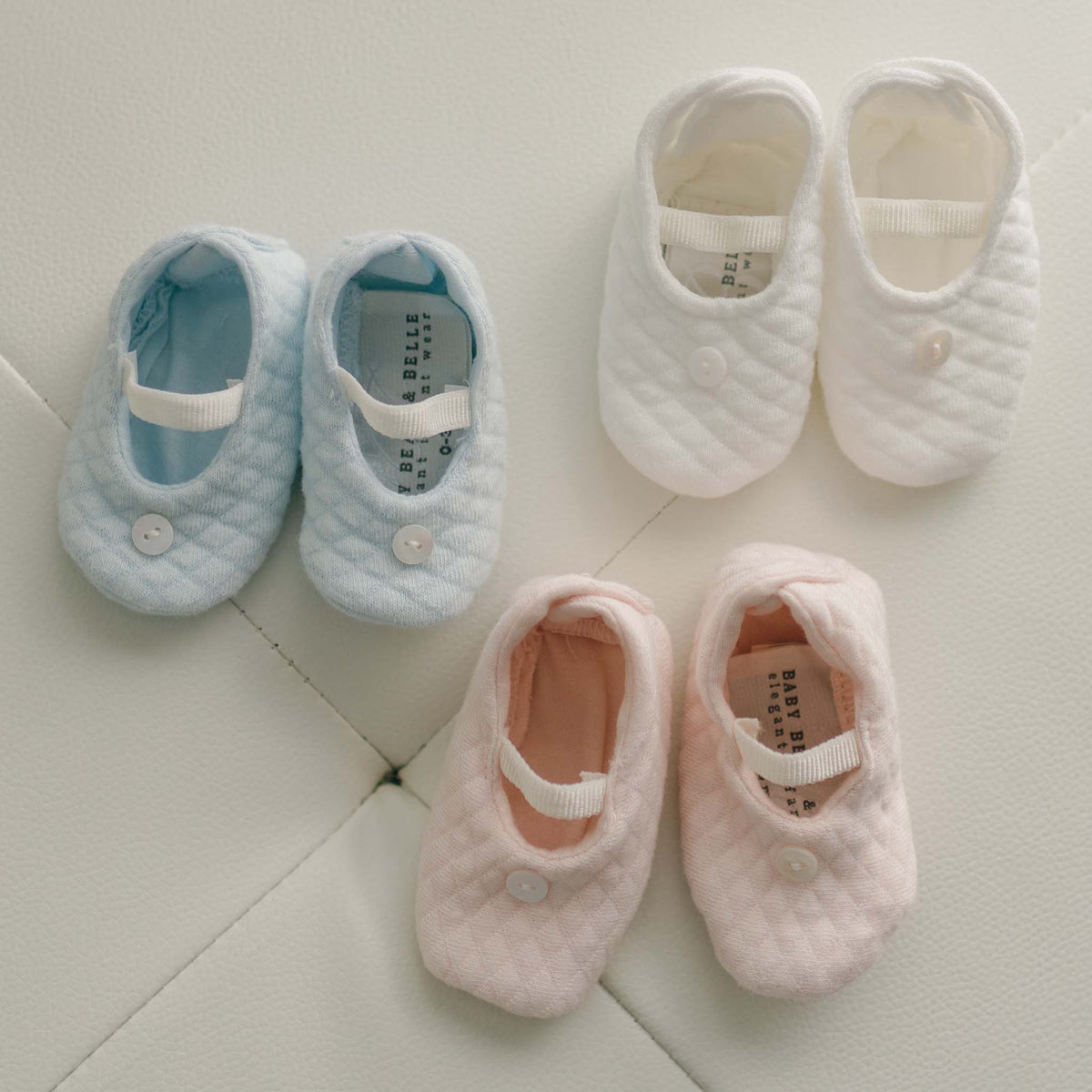Three pairs of quilted baby booties in light blue, white, and pale pink, arranged on a light textured surface. Each pair has a small button and a white elastic strap.