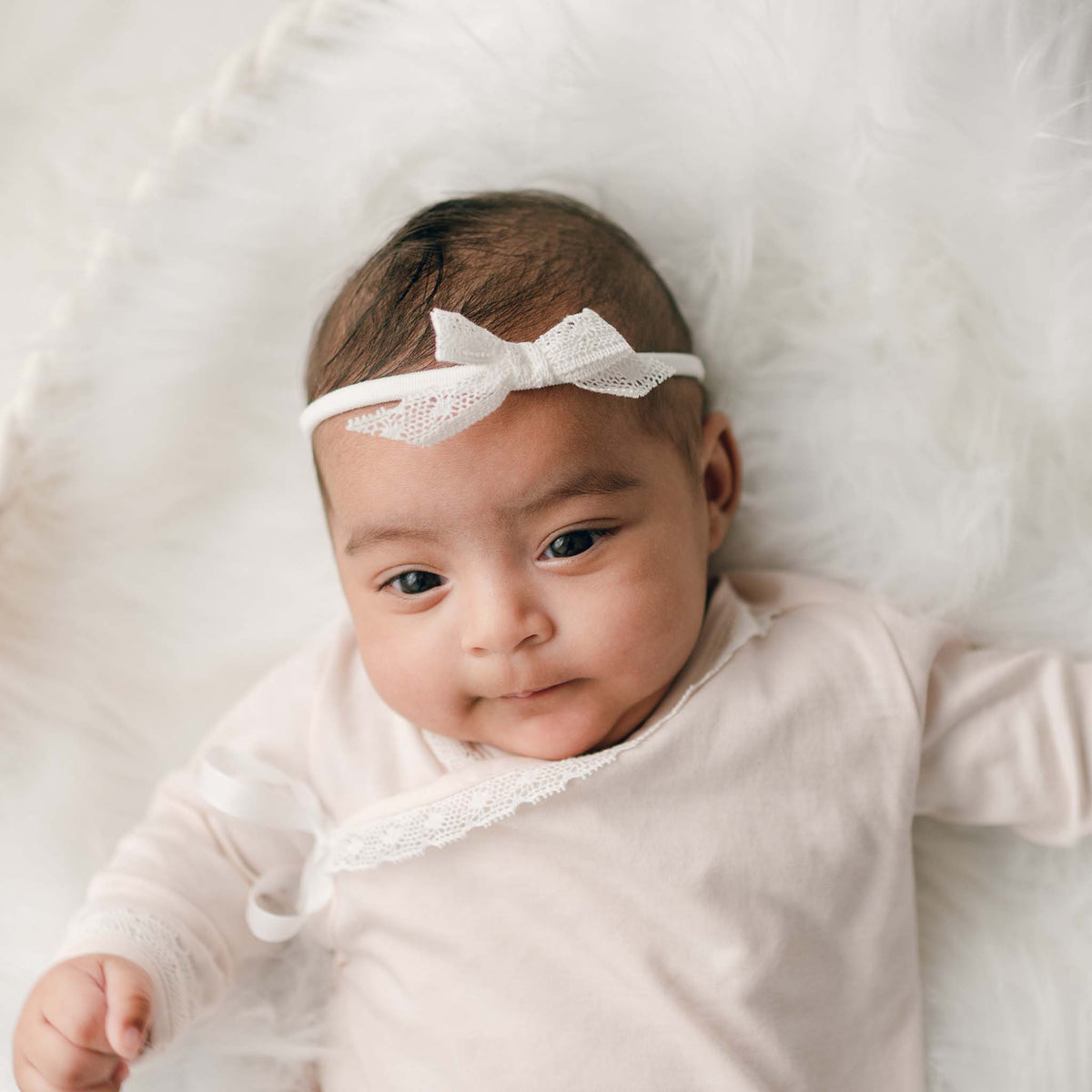 Baby boy in a pink layette and a lace bow headband laying on a white fur blanket