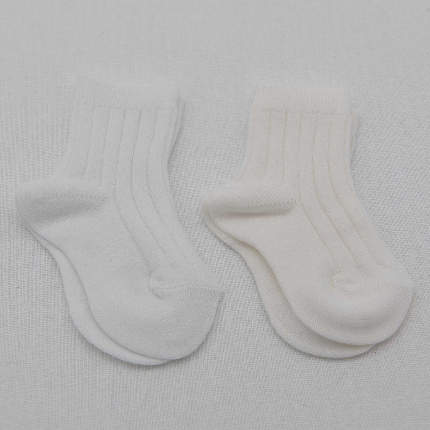 Two pairs of Ribbed Socks, one in a white cotton blend and the other in off-white, are displayed side by side on a light fabric surface. Both pairs are small and appear to be designed for an infant.