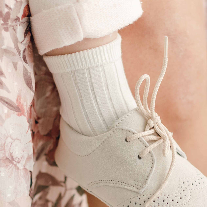 A close-up of a child's foot wearing a cream-colored lace-up shoe with perforated details. The child is also wearing soft Ribbed Socks made from a cotton blend, with the leg partially covered by rolled-up floral-patterned fabric.