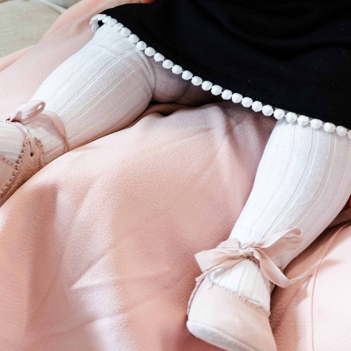 A close-up of a seated baby girl wearing stylish ribbed tights with ribbon ties at the ankles, pink pointed shoes.