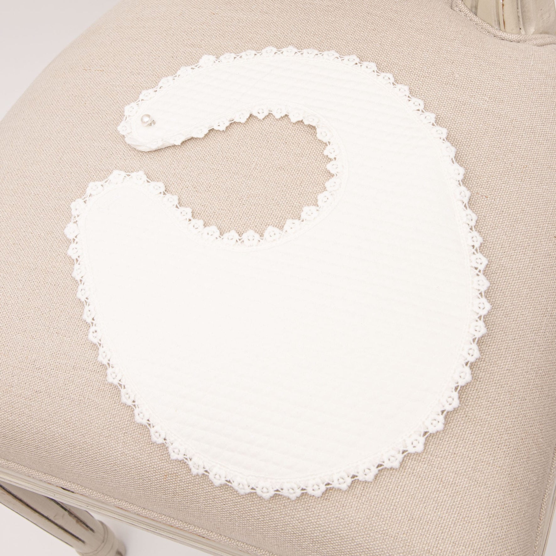 The Rose Accessory Bundle - Save 15% includes a white baby bib with a lace trim and matching lace baby shoes, elegantly displayed on a beige cushioned chair. The bib's scalloped edge and intricate detailing make it an exquisite addition to any baptism accessory set.