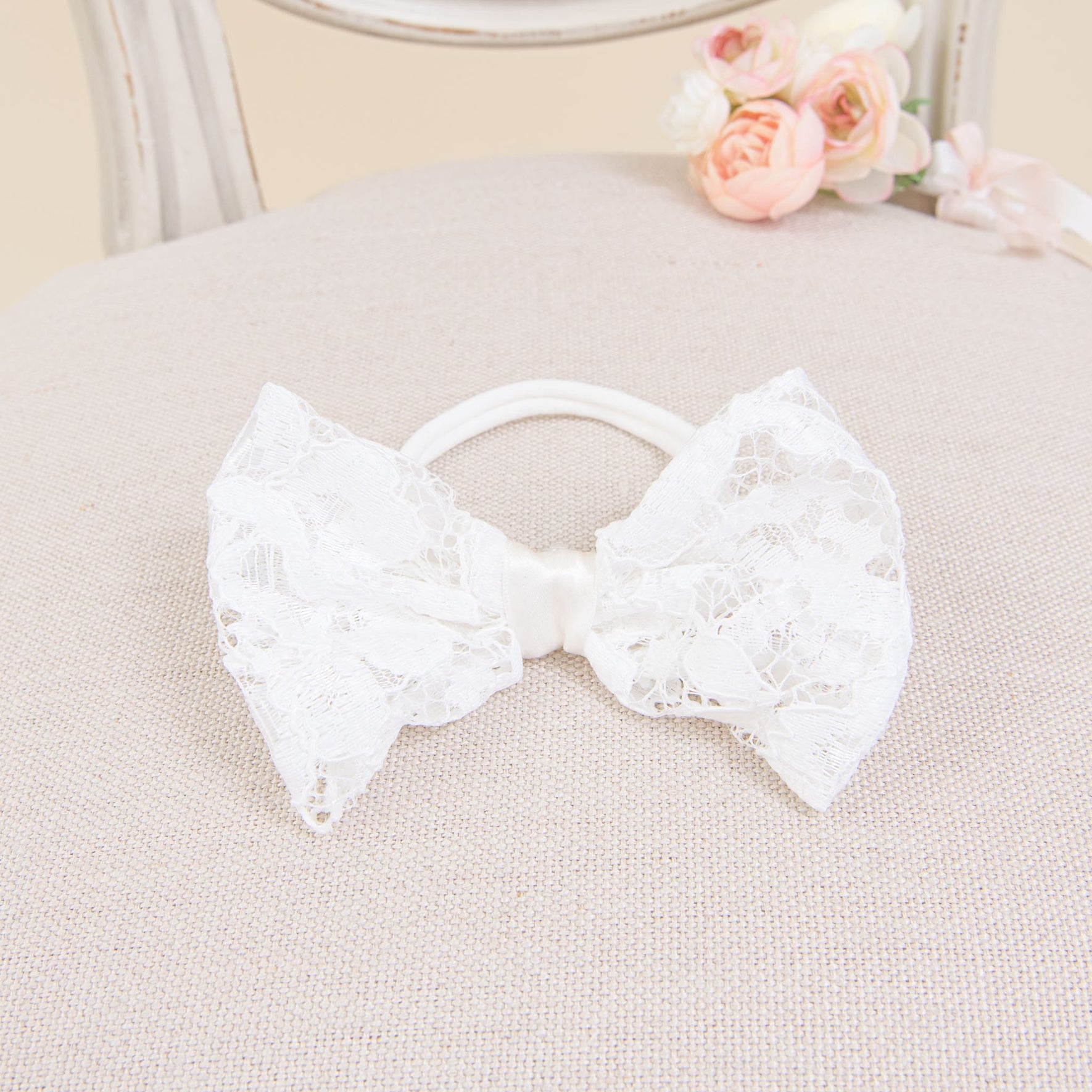 A white lace bow is set against a beige textured surface, reflecting the delicate charm found in lace baby shoes. In the background, a chair and a small bouquet of pink and white flowers can be seen slightly out of focus, ideal as elements of the Rose Accessory Bundle - Save 15%.