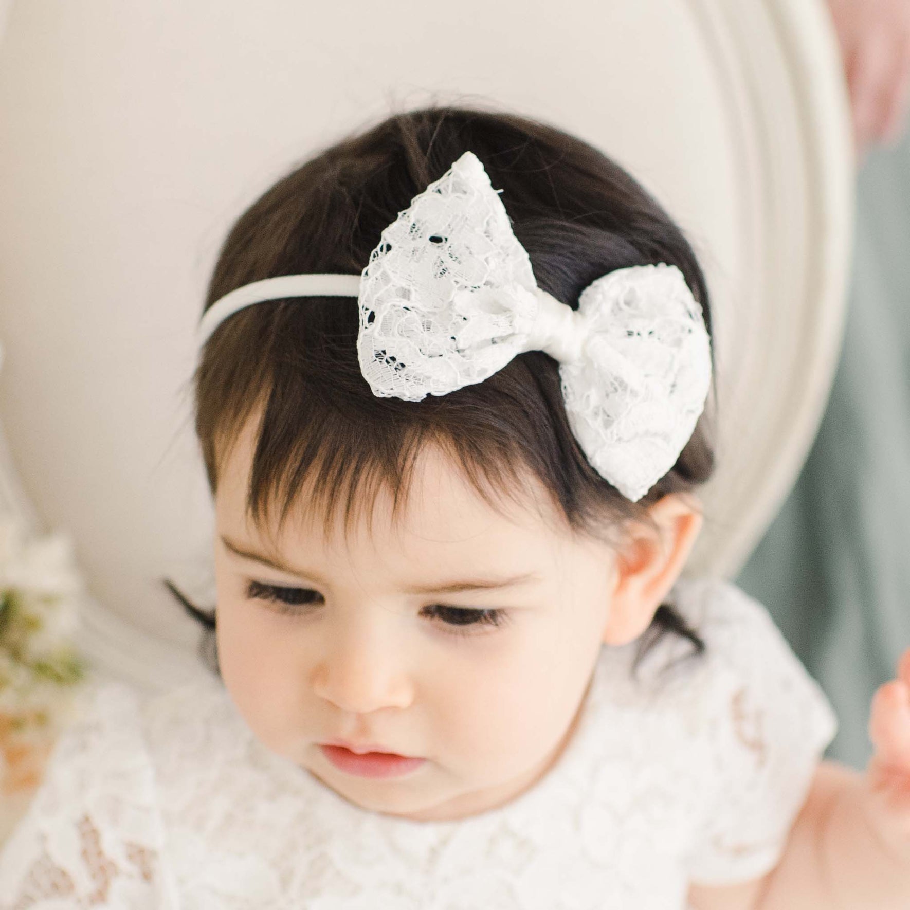 A toddler with dark hair wearing a white lace dress and a Rose Bow, looking down thoughtfully. The background is soft and neutral.