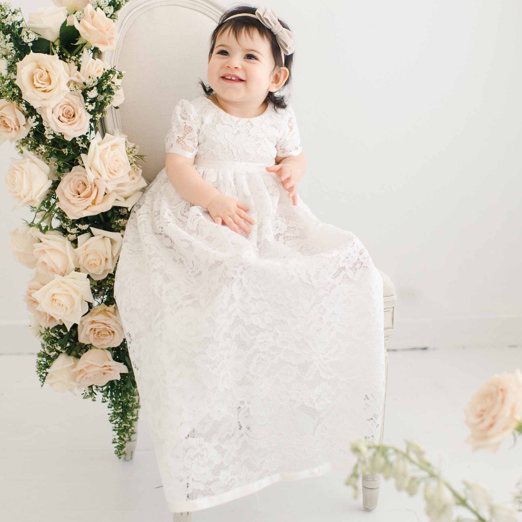 A toddler girl with a joyful smile, wearing an ivory lace Rose Christening Gown and Rose Velvet Bow Headband, sits on an elegant chair adorned with pink roses in a light, bright room.