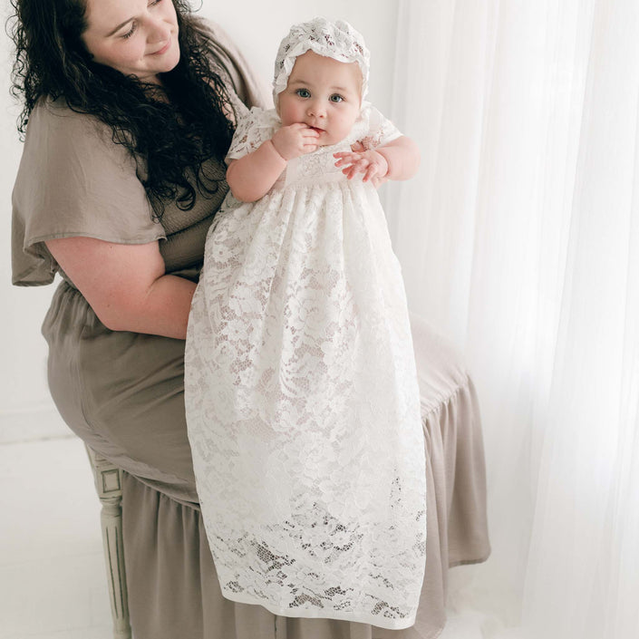 A mother holding her baby dressed in the Rose Christening Gown & Bonnet. The woman, smiling gently, gazes at the baby who appears curious, set against a bright, softly lit backdrop.