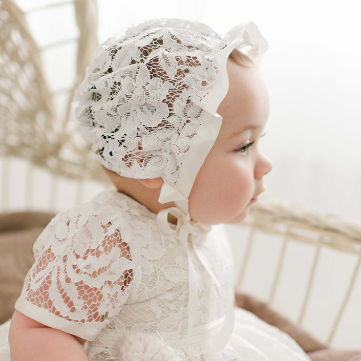 A baby in the Rose Lace Christening Bonnet and matching gown sits in a wicker chair, gazing thoughtfully to the side, showcasing the delicate lace details of the bonnet.