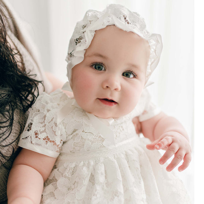 A close-up of a baby girl dressed in the Rose Lace Christening Bonnet and matching baptismal gown looks directly at the camera with an expression of curiosity.