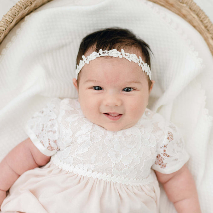 A baby wearing a handmade white lace dress and the Rose Lace Headband is lying on a soft, white blanket in a woven basket. The baby is smiling, and the setting appears to be comfortable and serene.