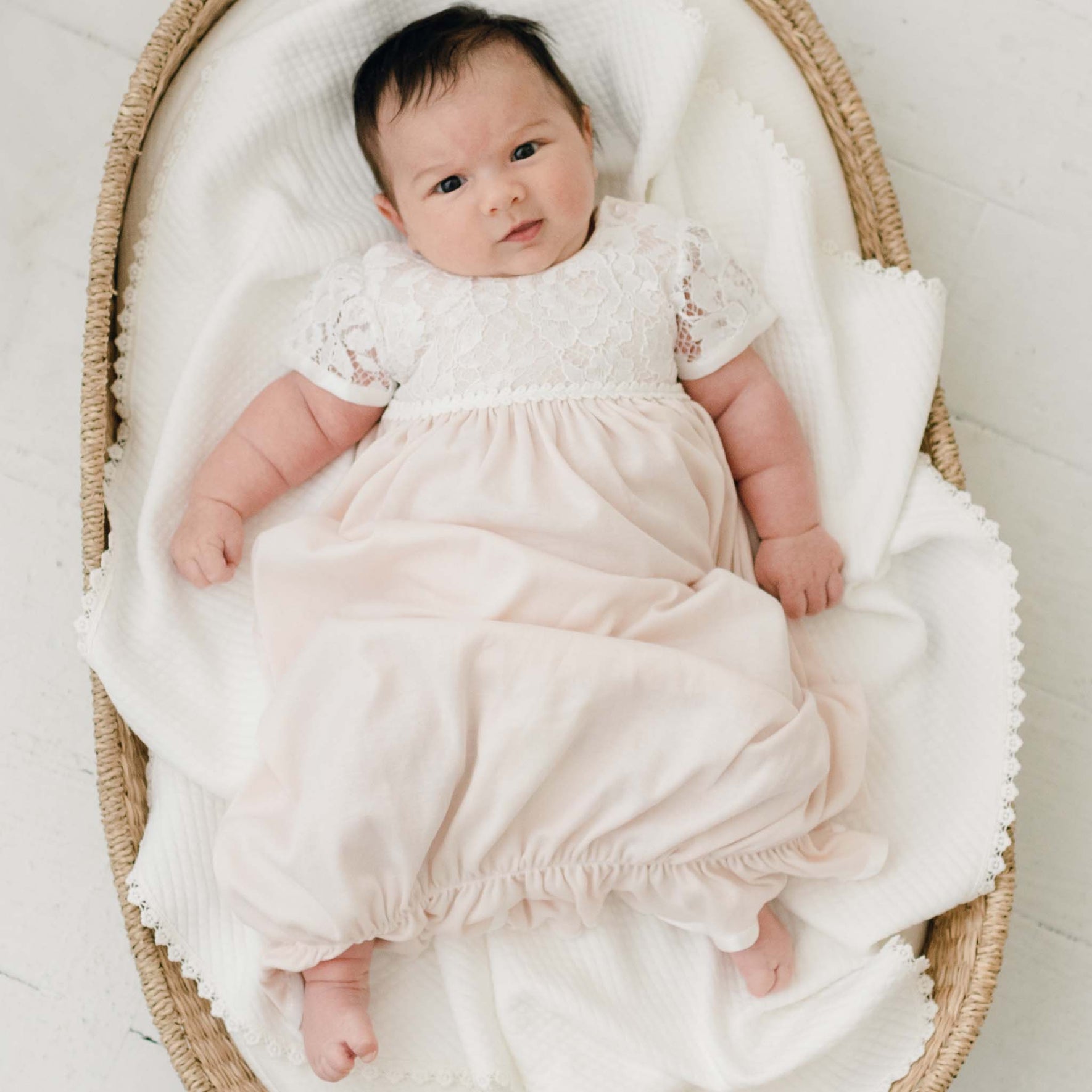 A baby with dark hair lies in a wicker bassinet, dressed in the Rose Layette & Bonnet. The baby is looking up with a neutral expression, surrounded by a white blanket. The bassinet rests on a light-colored surface.