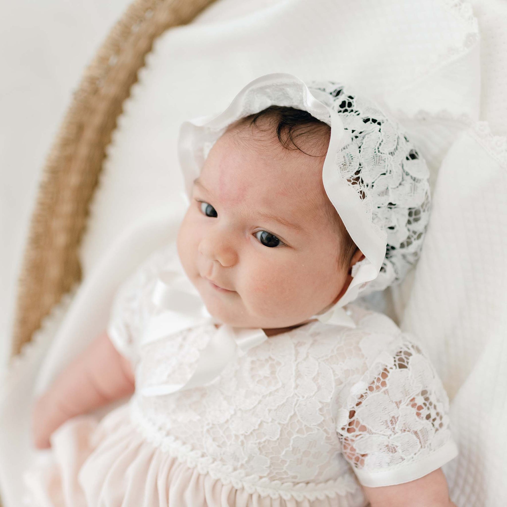 A baby wearing a Rose Layette & Bonnet is lying on a white blanket. The baby is looking slightly to the side with a calm expression. She is laying in a wicker bassinet.