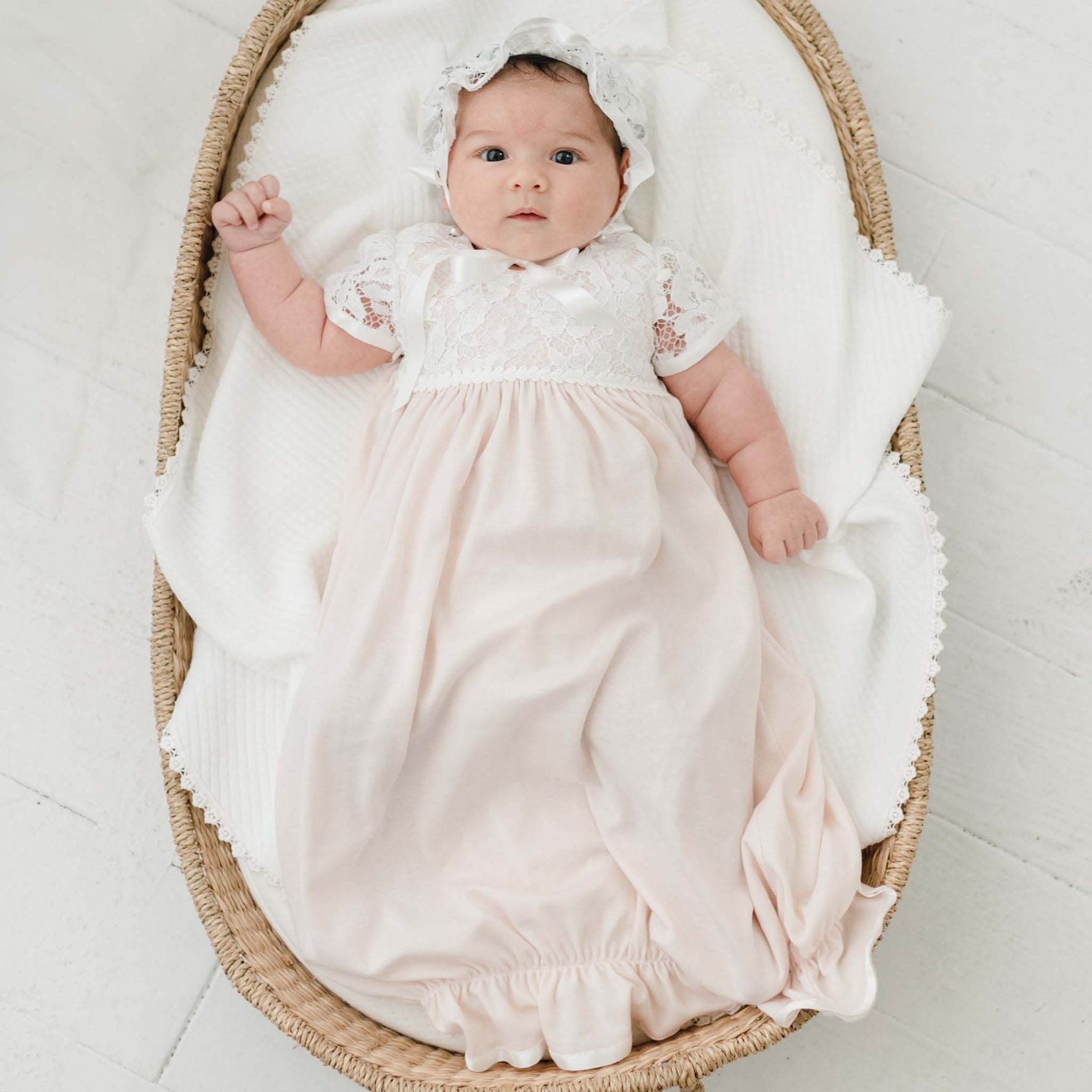 A baby lies in a woven basket on a white blanket. She is wearing the Rose Layette & Bonnet. The baby's right arm is slightly raised, and their eyes are open, looking directly at the camera.