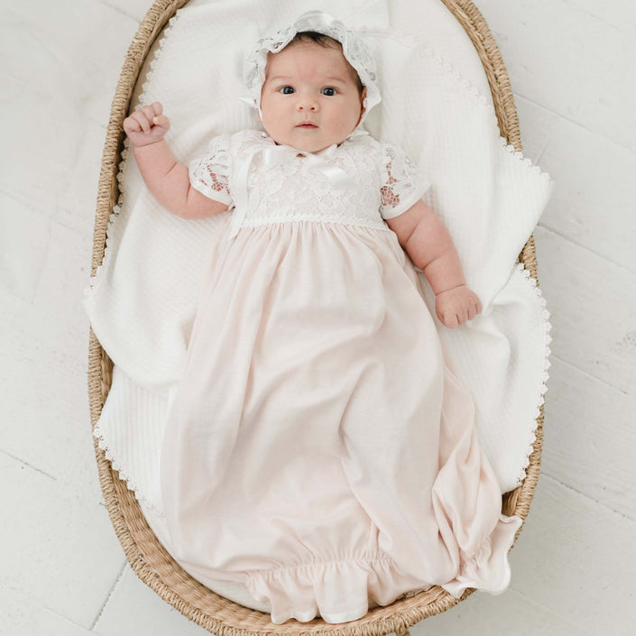 A baby lies in a woven basket on a white blanket. She is wearing the Rose Layette & Bonnet. The baby's right arm is slightly raised, and their eyes are open, looking directly at the camera.