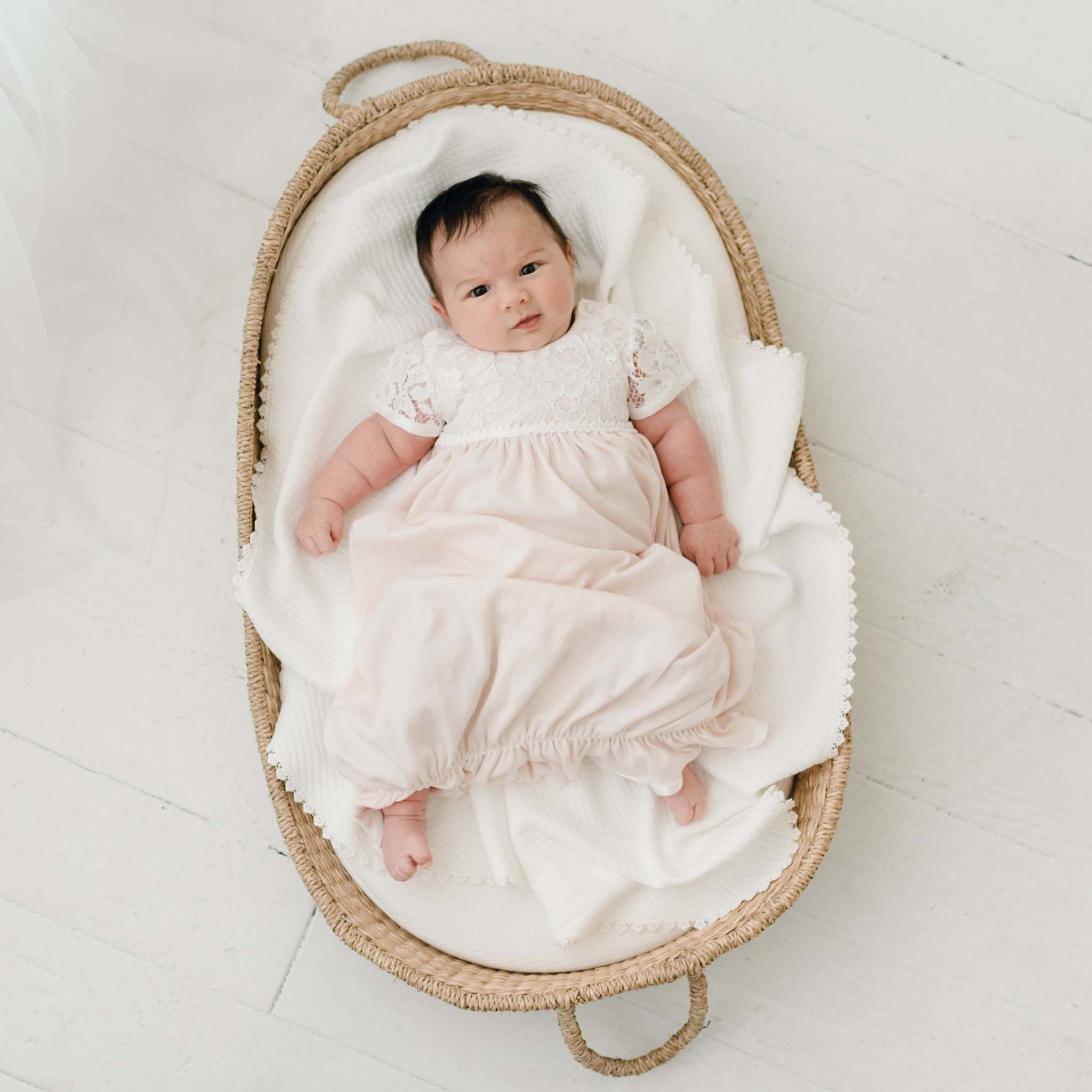 A baby dressed in a Rose Layette & Bonnet is lying in a wicker basket lined with a white blanket. The basket rests on a white wooden floor, and the baby gazes up with a neutral expression.