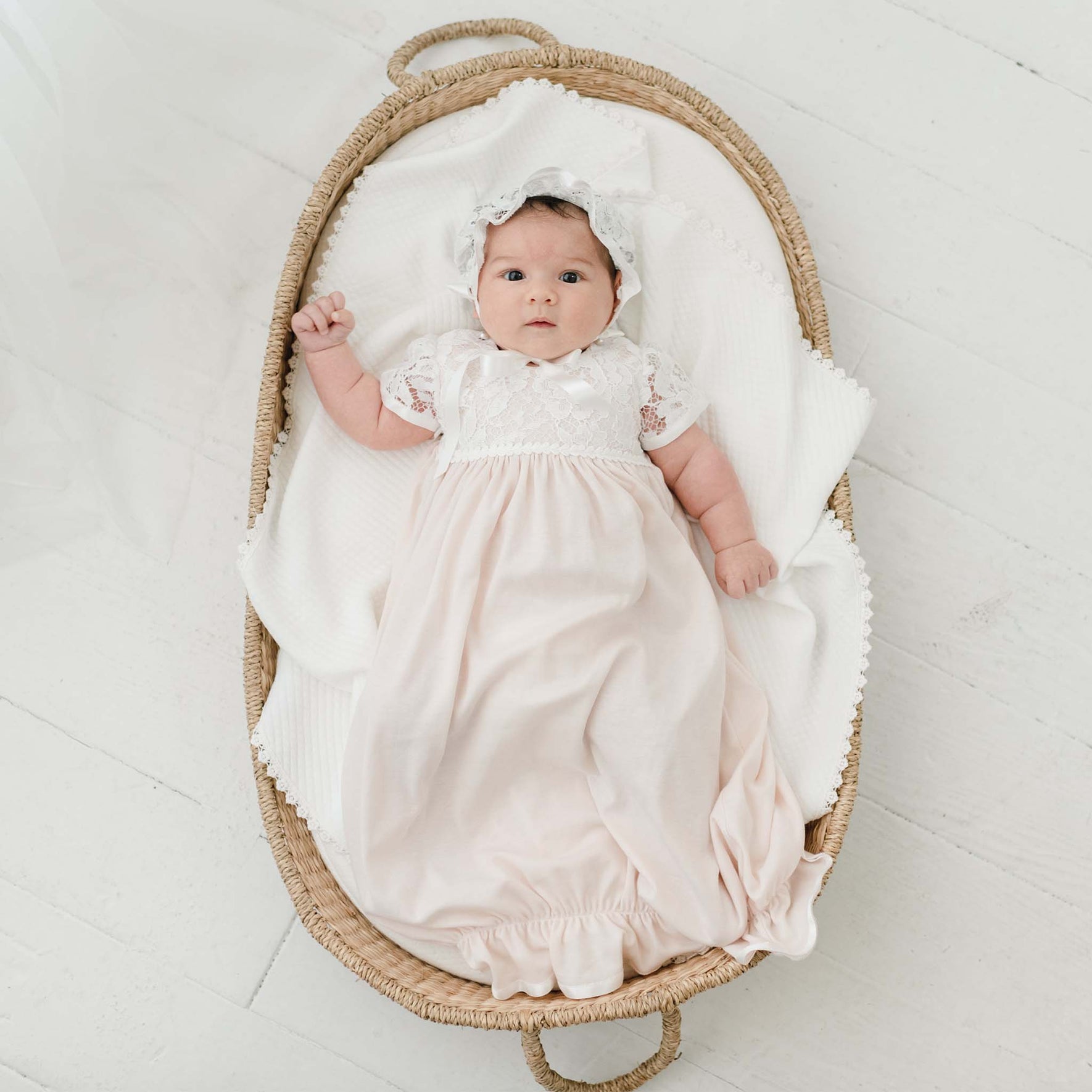 A baby is lying in a woven basket on a white blanket, swathed in a long white dress and a delicate Rose Layette & Bonnet. With one arm slightly raised, the newborn layette exudes charm against the light-colored floor backdrop.