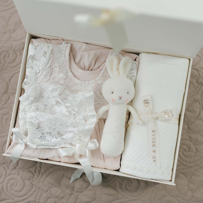 A white box contains a folded pink dress with rose patterned lace overlay, a white plush rabbit toy with a stitched face, and a pima cotton quilted blanket tied with a ribbon labeled "Rose Newborn Gift Set - Save 10%." The items are neatly arranged on a quilted beige surface.
