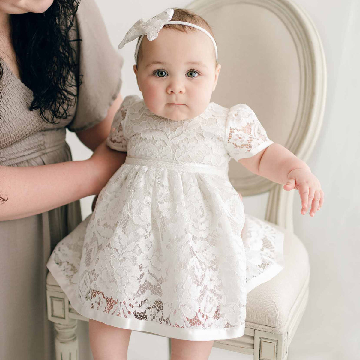 A baby in an ivory lace dress and headband sits on a cream chair. An adult, partially visible, supports the baby. The setting has soft lighting and neutral tones.