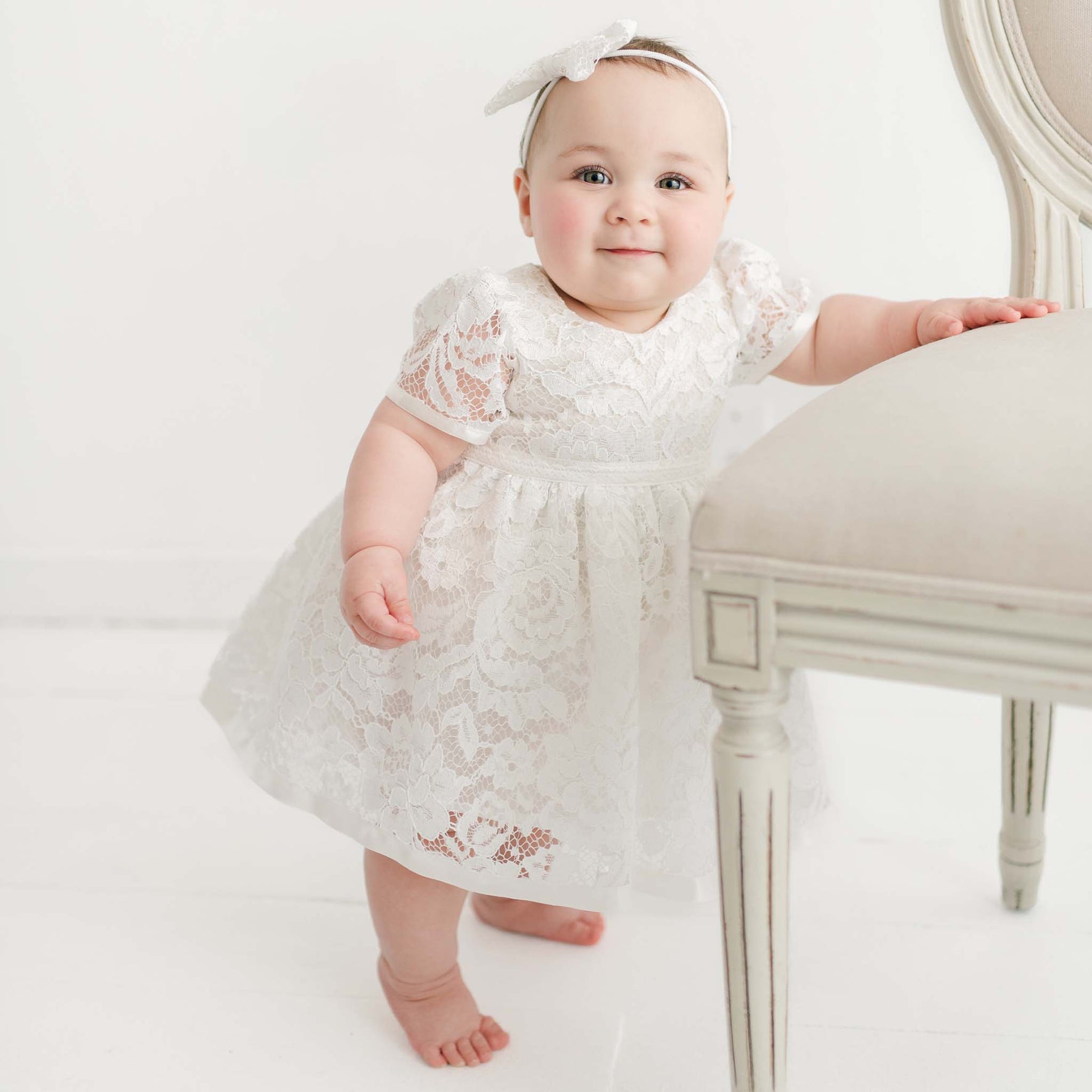 A baby girl in the Rose Romper Dress and matching Bow Headband stands barefoot, leaning on a vintage chair in a bright, white studio setting.