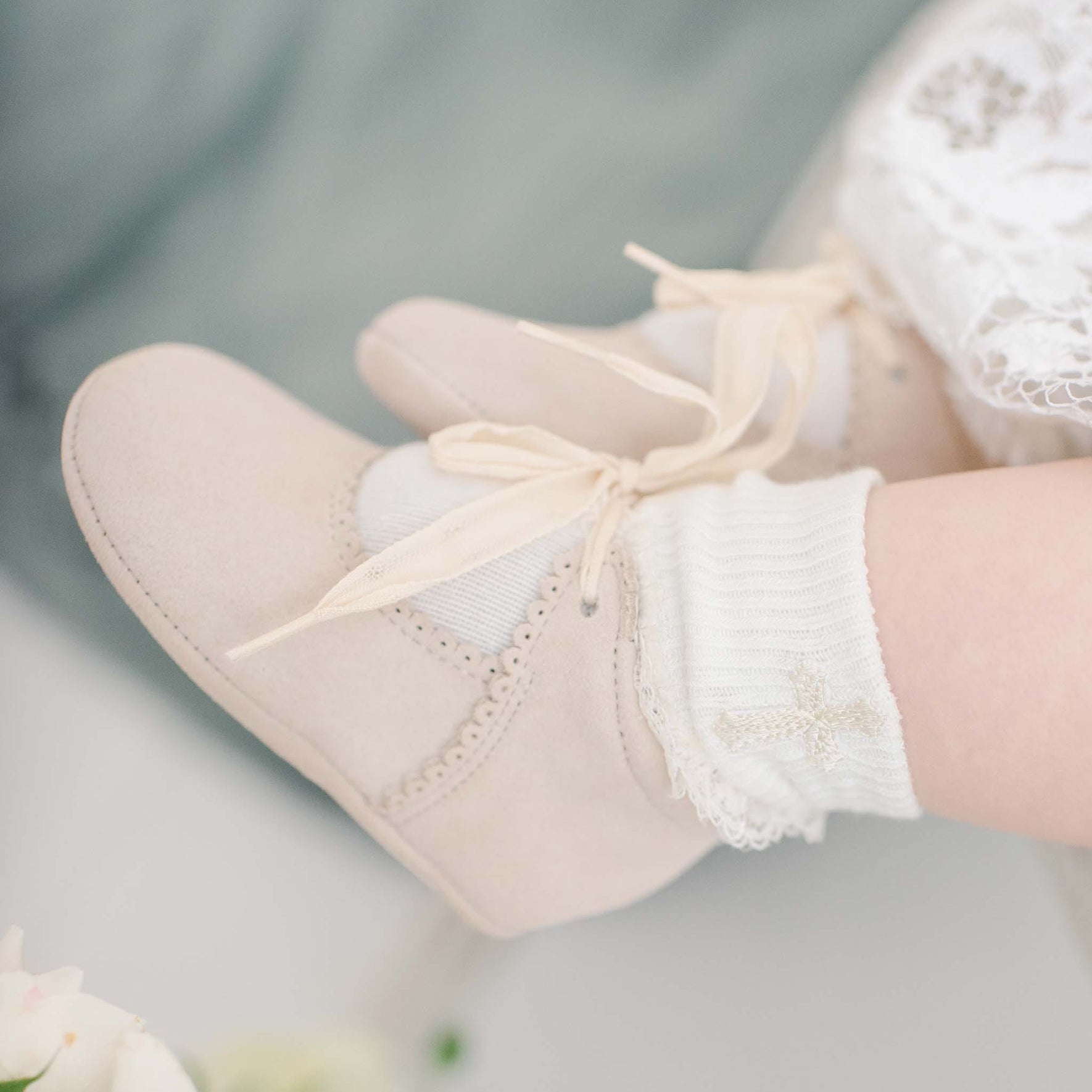 Close-up of a baby wearing ivory lace-trimmed socks and Rose Suede Tie Mary Janes with ribbon ties. The baby, in Baby Beau & Belle white lace, rests on a soft surface with blurred floral elements in the corner.