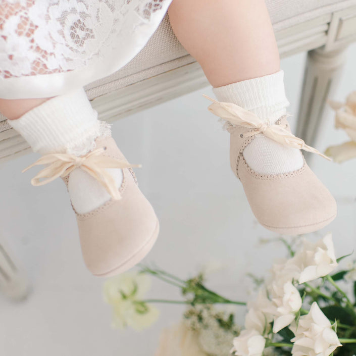 A baby sits on a bench wearing cream-colored Rose Suede Tie Mary Janes adorned with delicate bows. The suede leather shoes are paired with white socks, complementing the baby's white lace outfit, while flowers are elegantly arranged underneath.