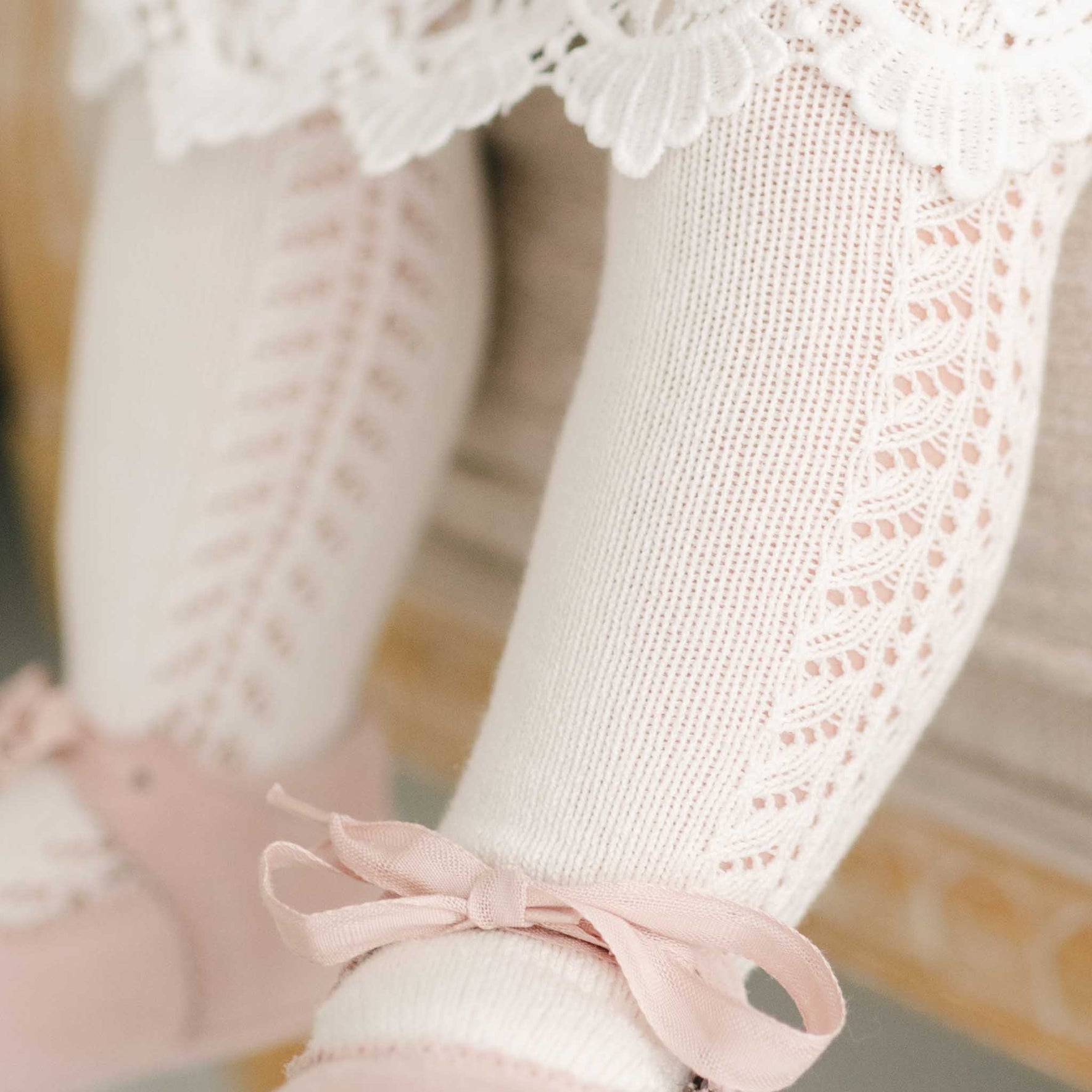 Close-up of a child's legs wearing the Side Openwork Tights in cream, paired with soft pink shoes adorned with bows. The luxurious lace details on the tights and the textured design of the shoes exude a gentle, delicate style that enhances the entire outfit.