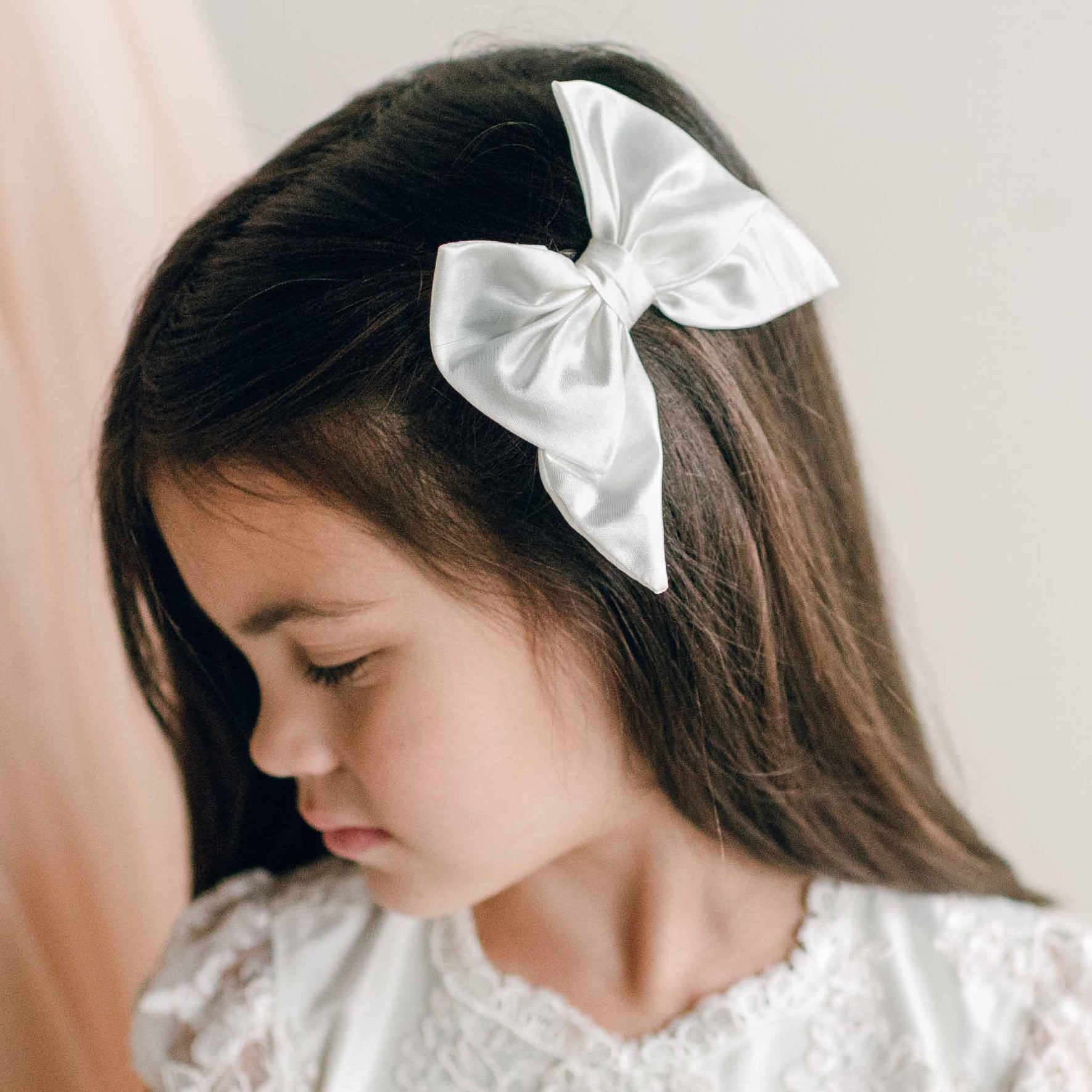 A young girl with long dark hair wears a white silk bow clip on the side of her head, complementing her lace-trimmed dress.