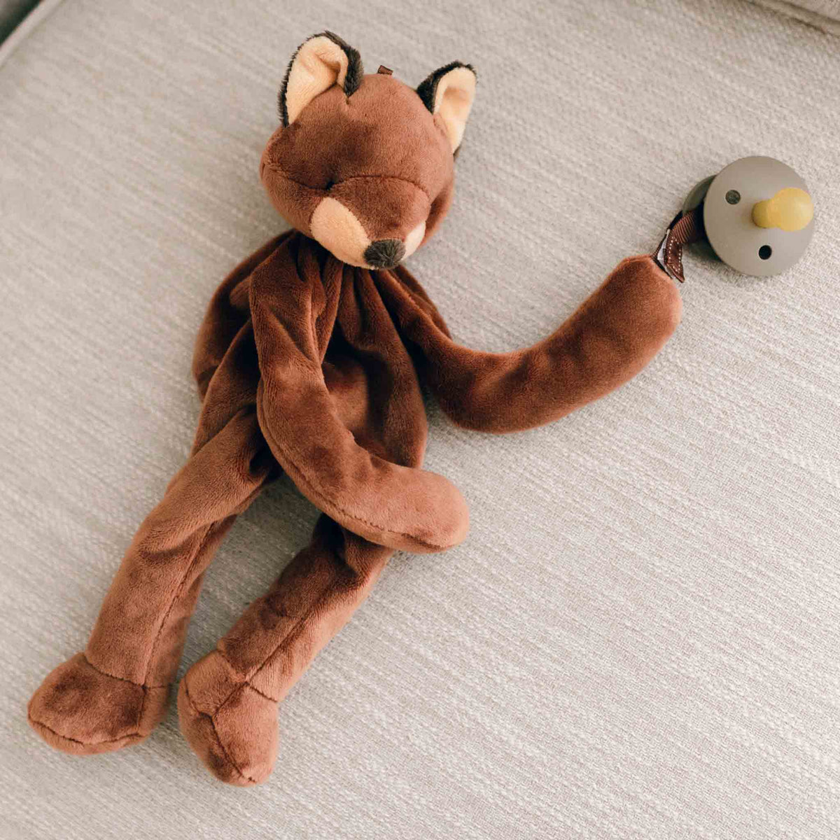 A plush Silly Fox Buddy, serving as a pacifier holder, rests on a light-colored fabric surface with one arm outstretched, holding a small gray and yellow rattle—making it the perfect baby gift.