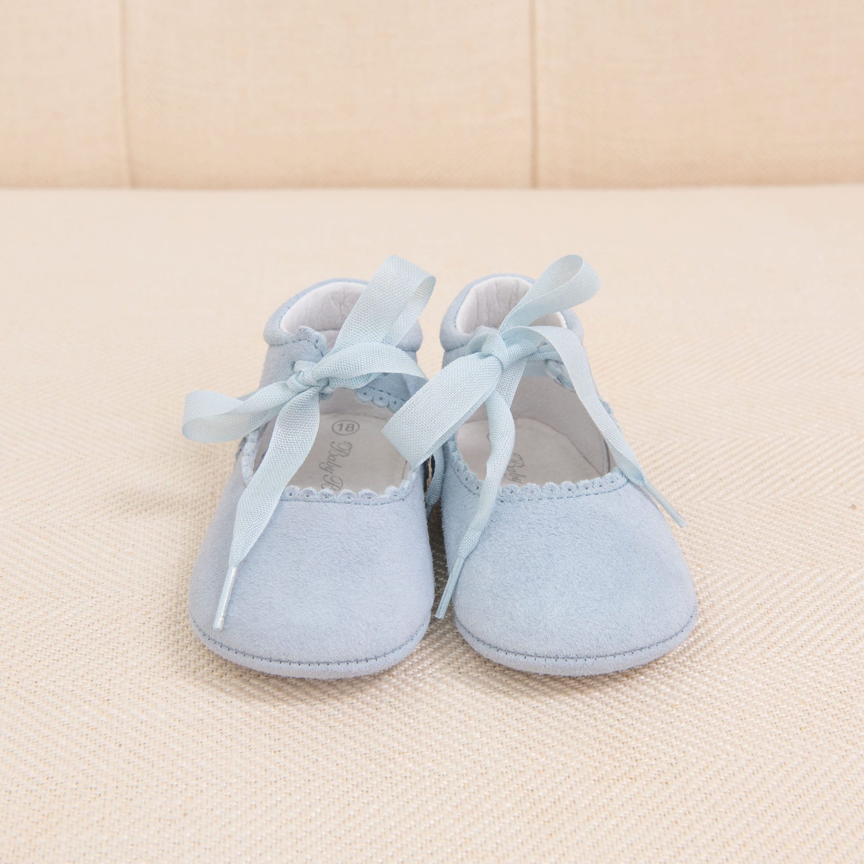 A pair of Sky Blue Suede Tie Mary Janes with ribbon laces is placed on a cream-colored textured surface. The handmade baby shoes have a suede-like texture and delicate scalloped edging along the opening.