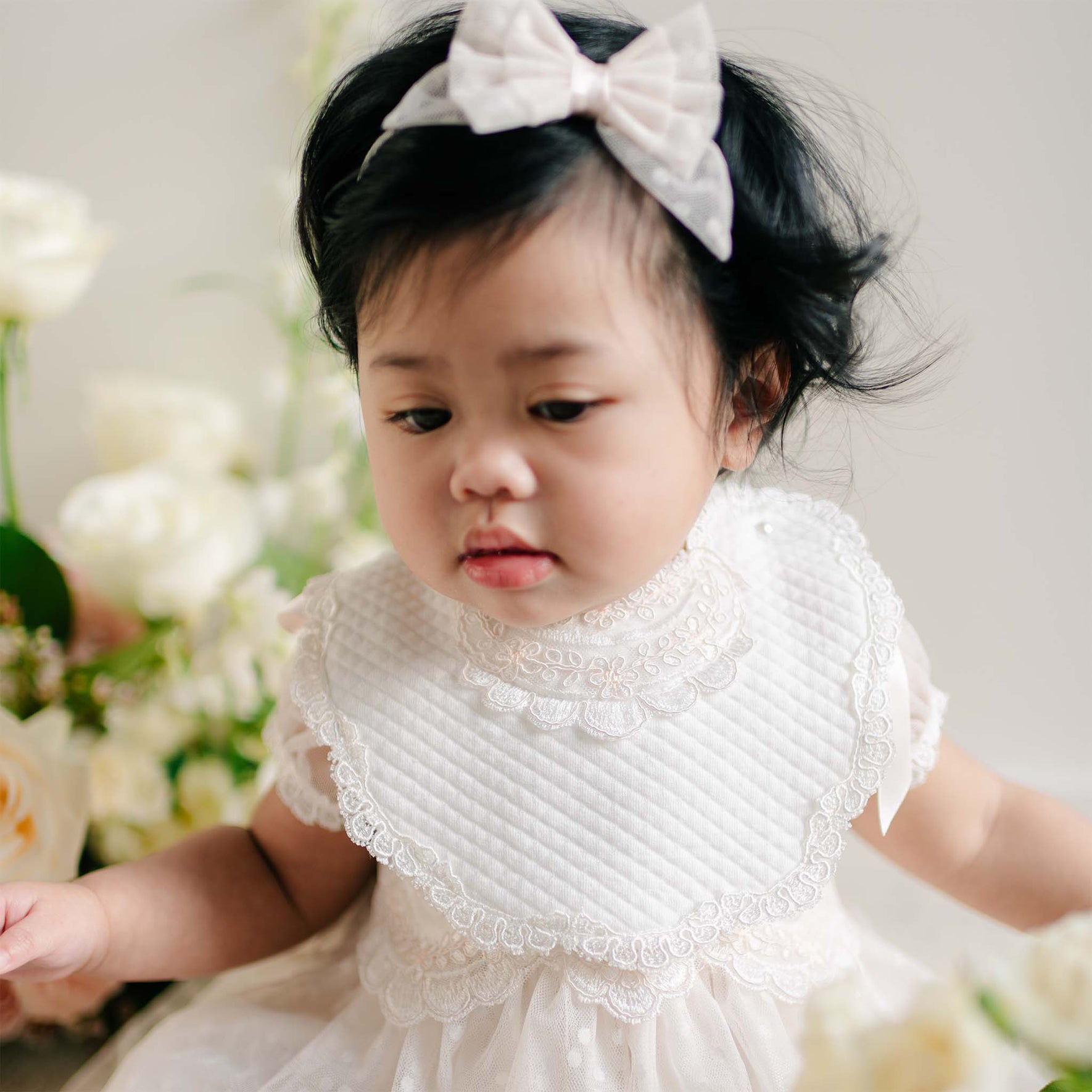 Baby girl wearing the Sophia Bib with scalloped lace trim over a white embroidered dress.
