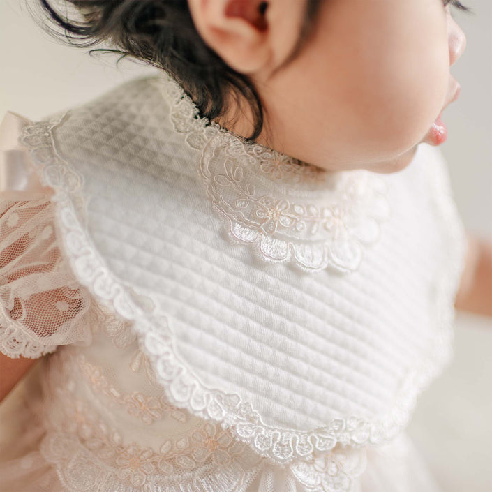 A close-up of the Sophia Bib, featuring a quilted white fabric with delicate lace trim and floral embroidery, worn over a coordinating lace dress.