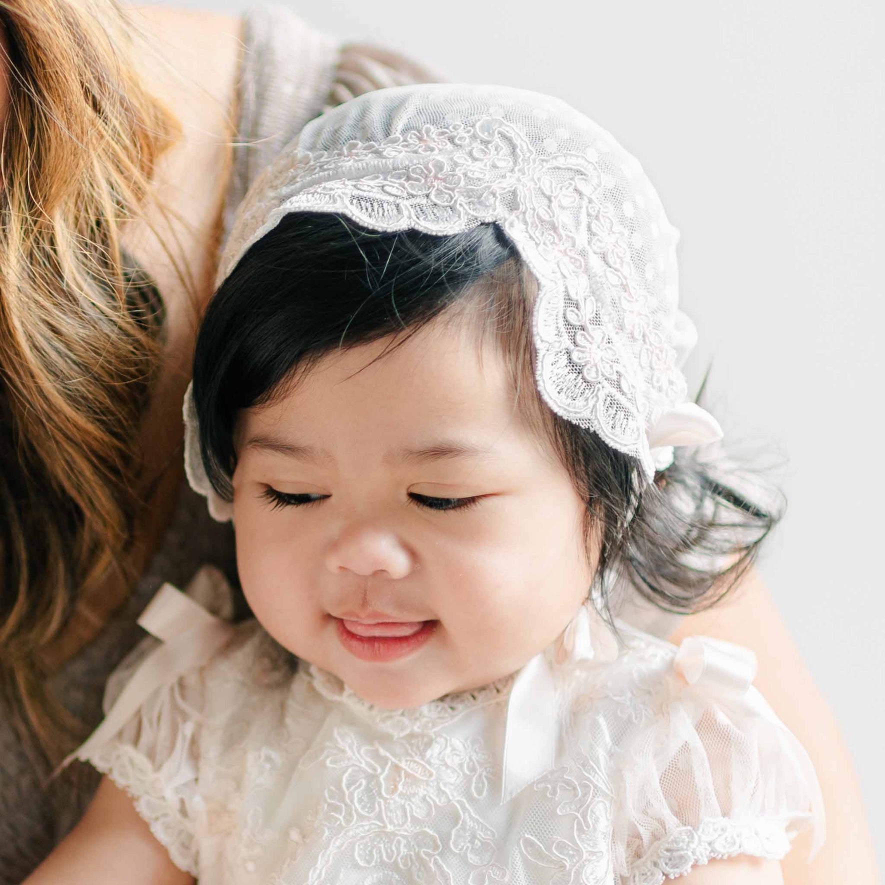A baby in the Sophia Bonnet and matching Sophia Dress, smiling with eyes closed, showing the detailed lacework and scalloped edges of the bonnet.