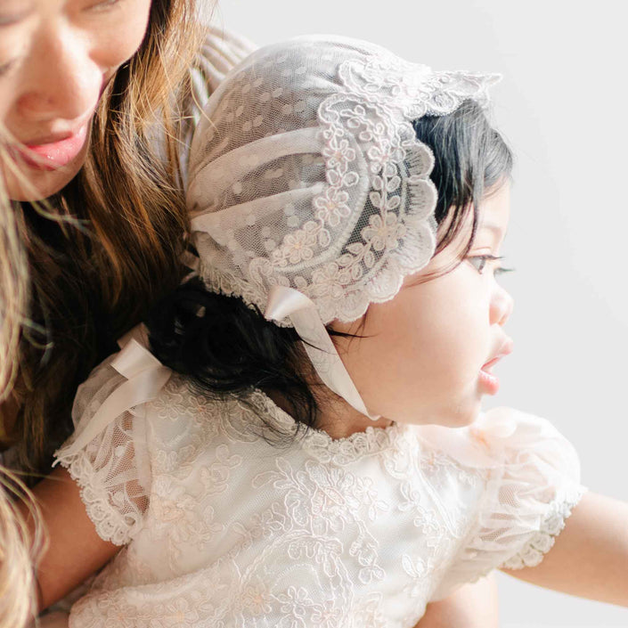 A close-up of a baby wearing the Sophia Bonnet, featuring delicate lace with floral embroidery and soft satin ribbon ties, paired with the Sophia Dress.