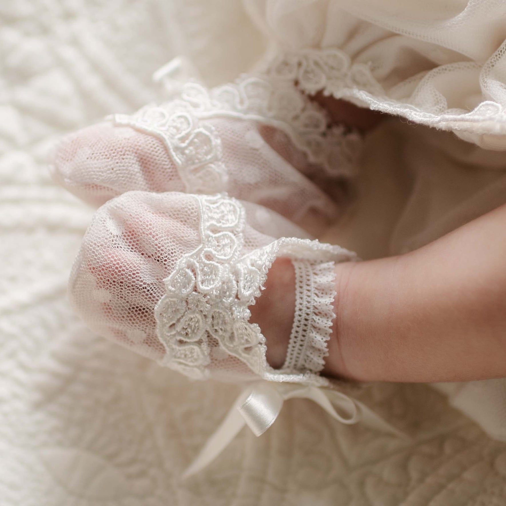 A baby's feet resting on a quilted surface, wearing the Sophia Booties with lace trim and a soft, sheer design.