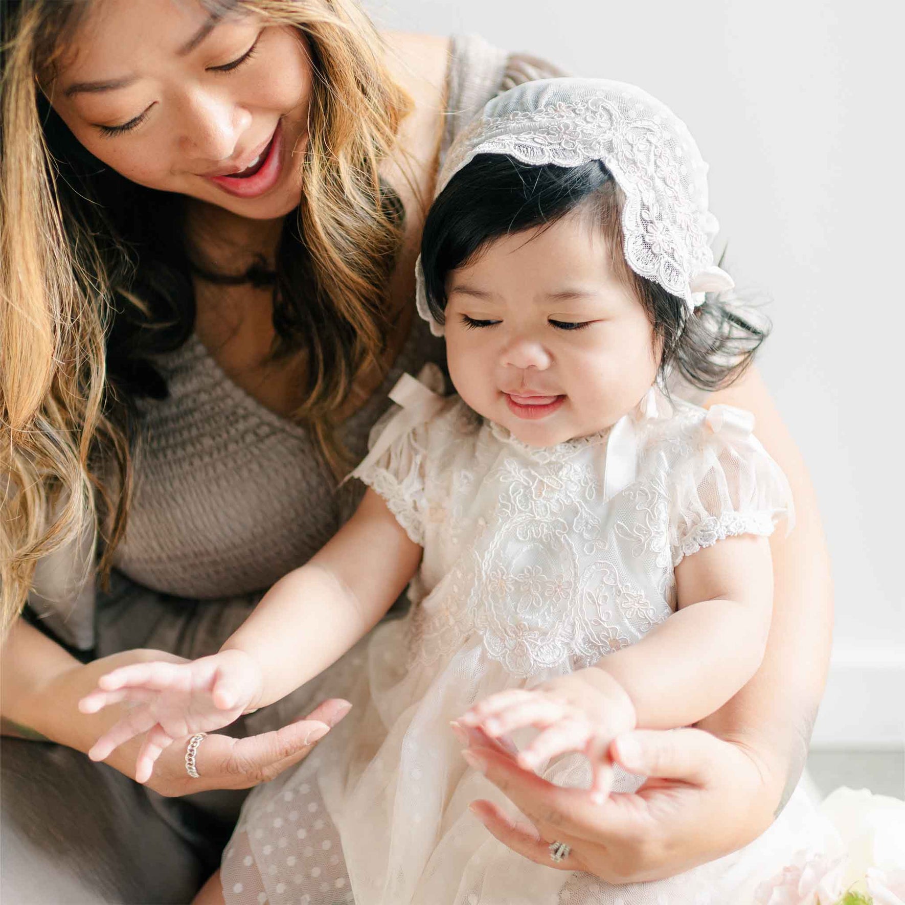 A mother holding her baby girl dressed in the Sophia Dress and matching bonnet, as the baby reaches forward with a joyful expression.
