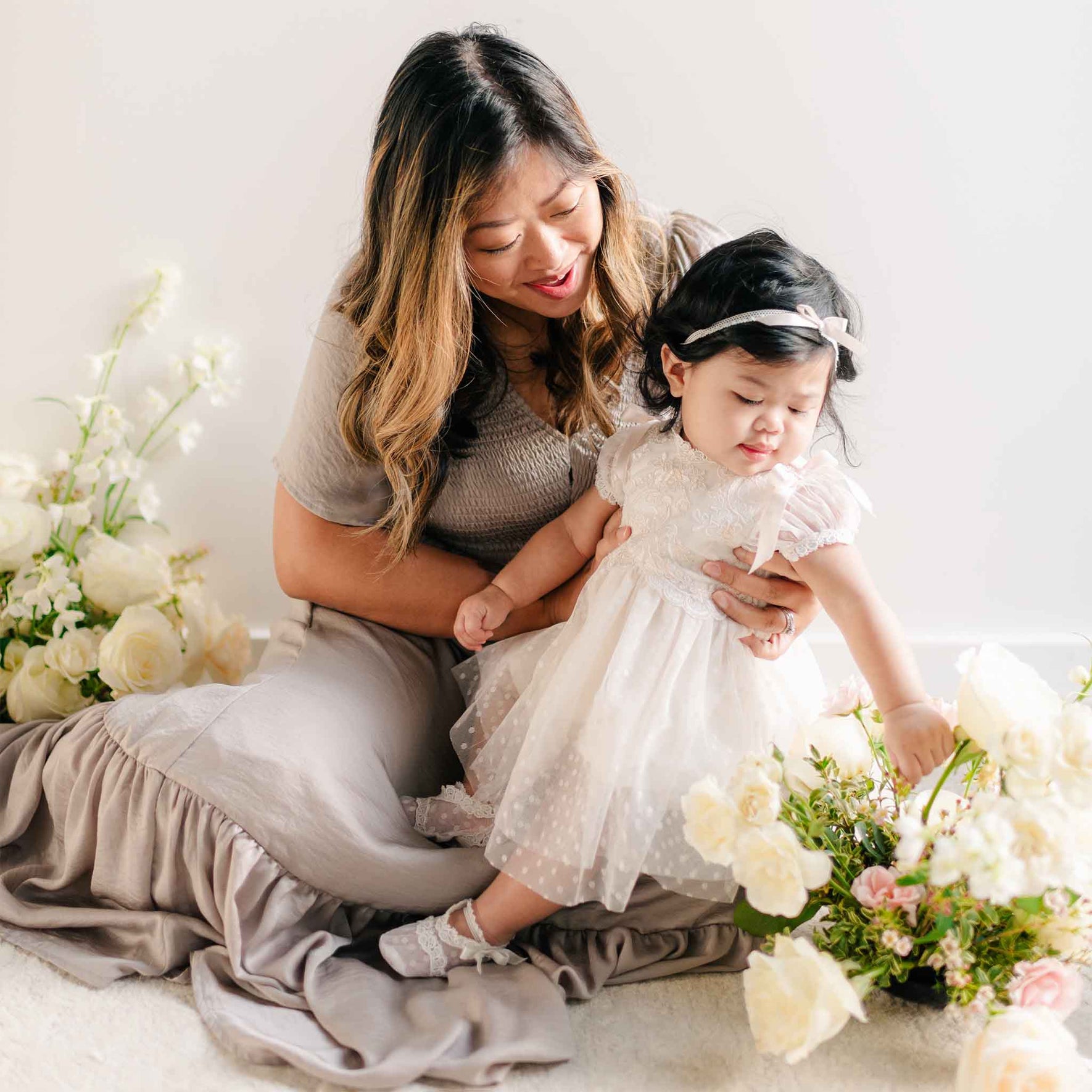A mother holding her baby girl in the Sophia Dress, with the baby reaching out to touch a bouquet of fresh flowers.