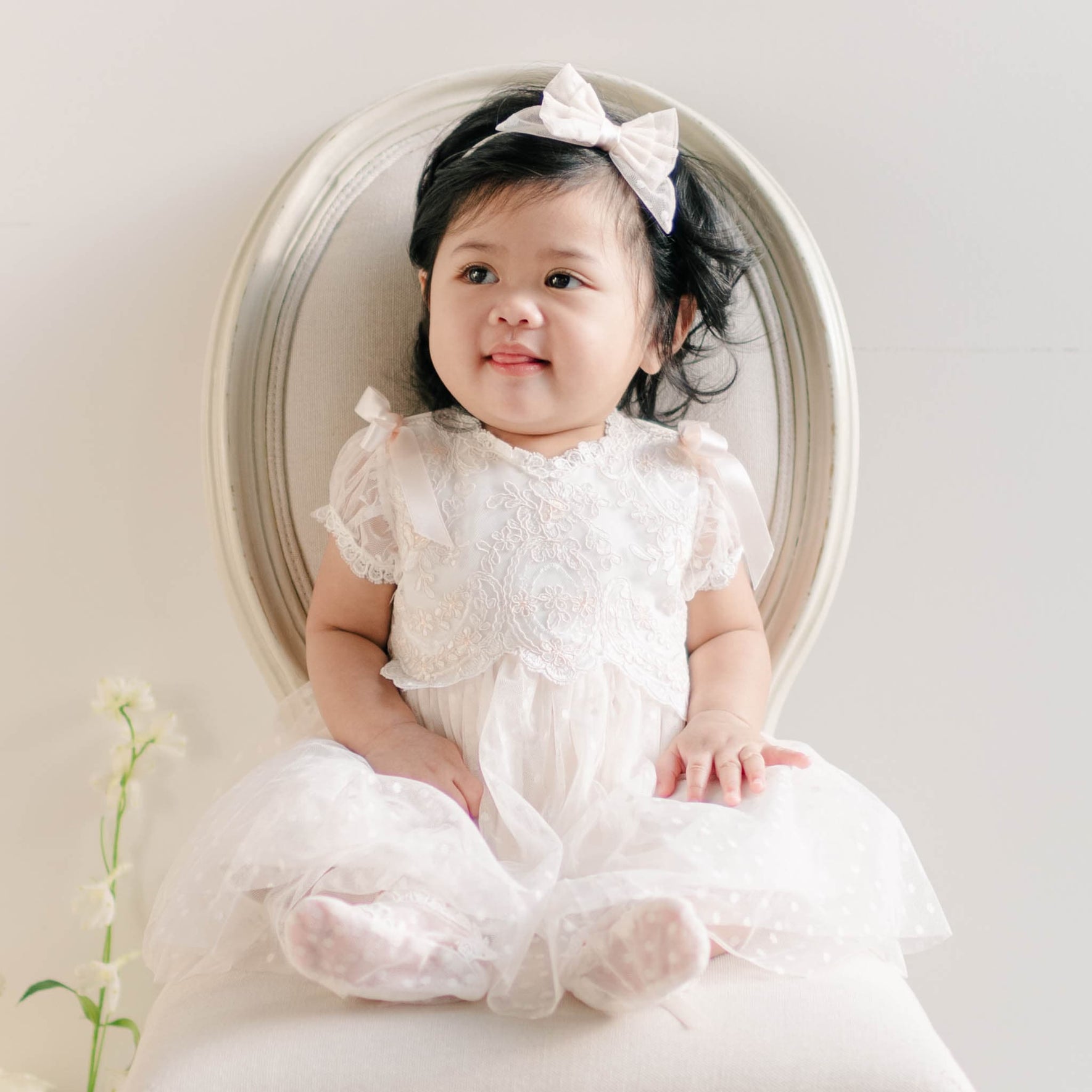 A baby girl sitting on an elegant white chair, dressed in the Sophia Dress with puffed sleeves and ribbon details, smiling softly.