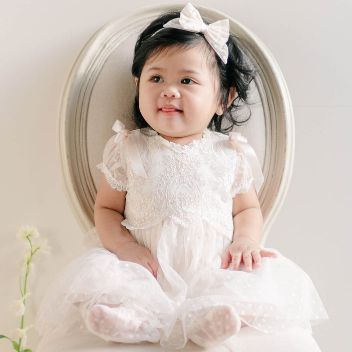 A baby girl sitting on an elegant white chair, dressed in the Sophia Dress with puffed sleeves and ribbon details, smiling softly.