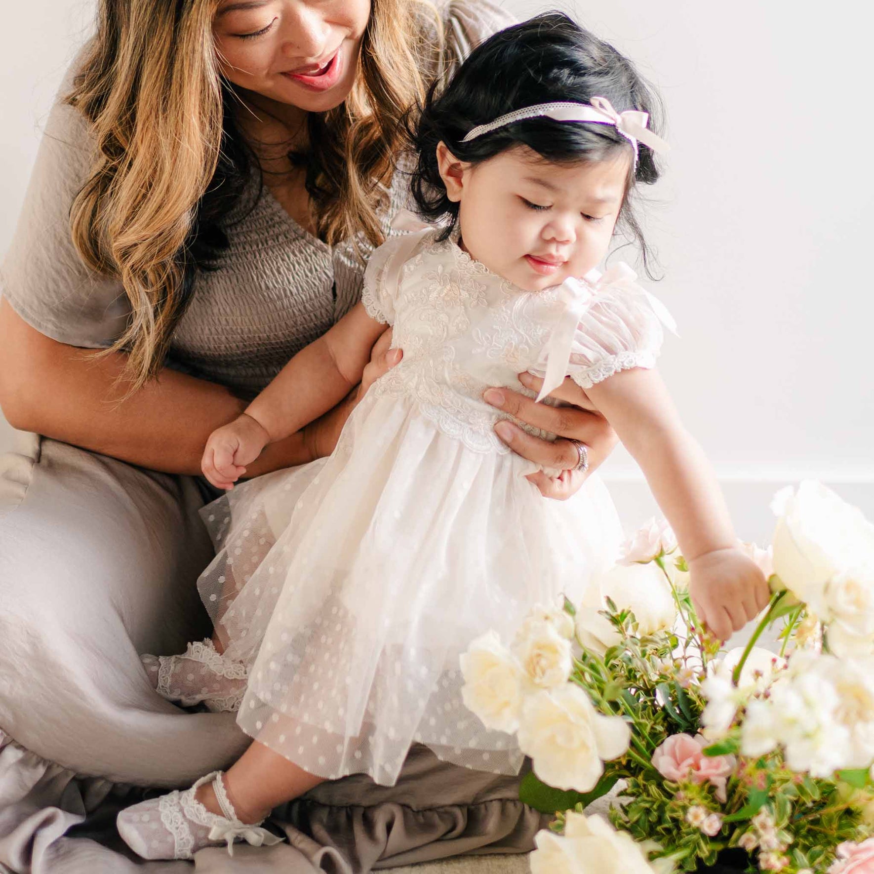 A baby girl in the Sophia Dress with puffed sleeves and lace details, reaching out to touch flowers while sitting on her mother’s lap.