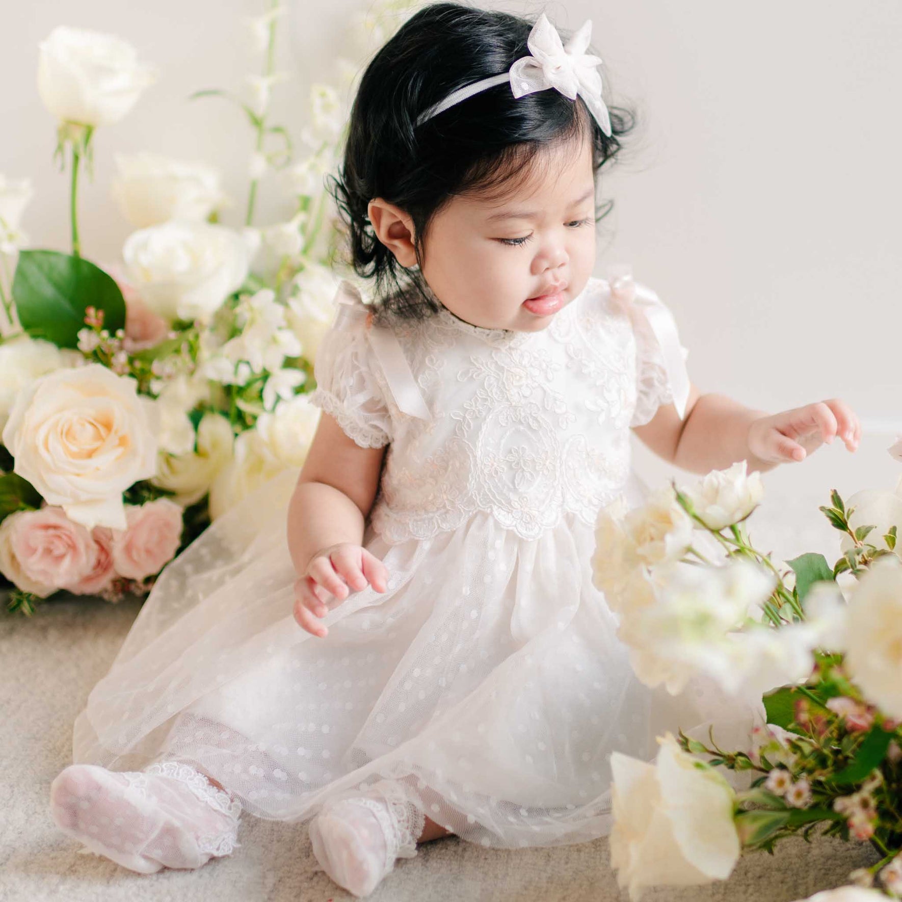 A baby girl in the Sophia Dress with satin ribbons on the sleeves, sitting among an arrangement of cream and blush roses.