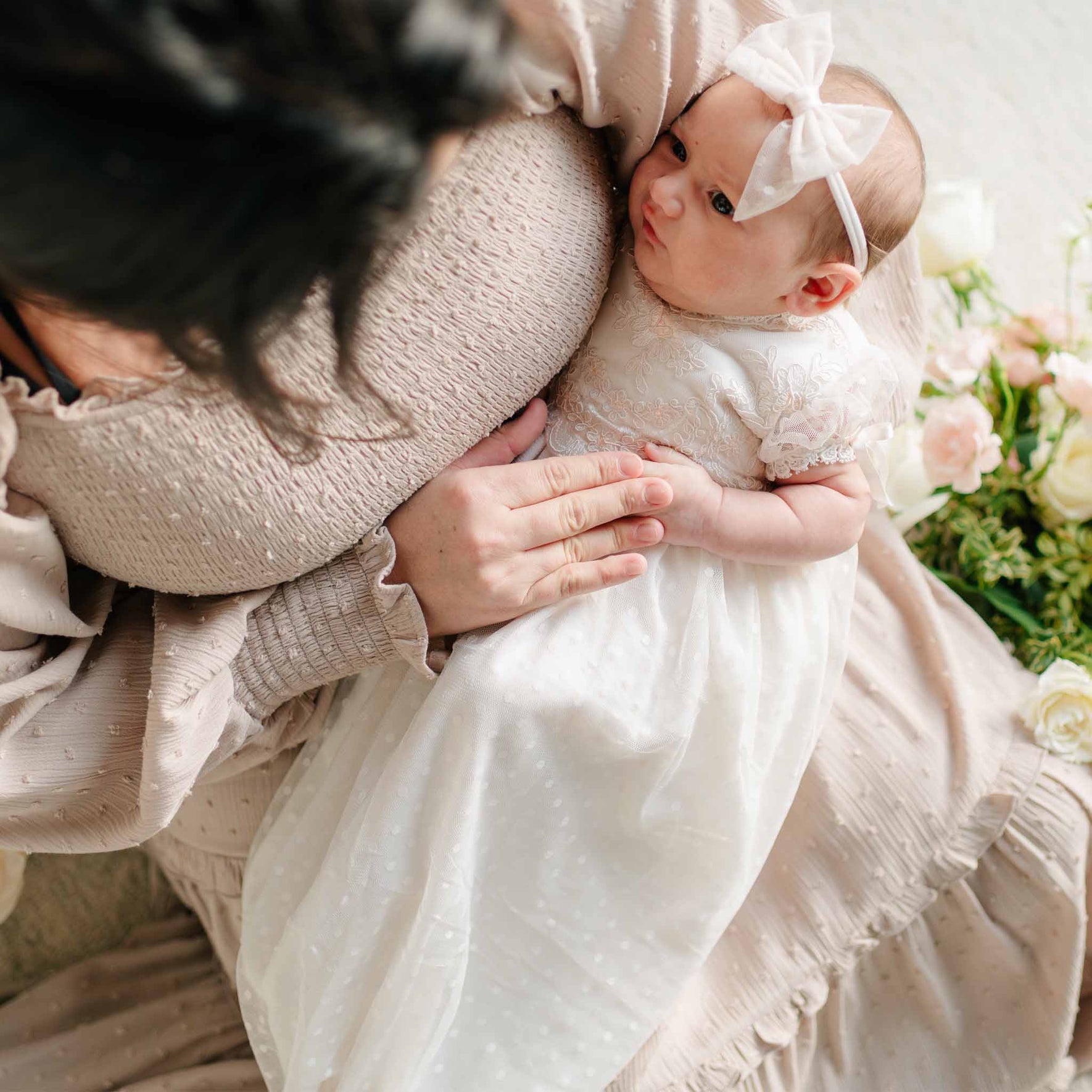 A newborn rests in their mother's arms, wearing the Sophia Lace Bow Headband along with a lace gown, gazing upward.