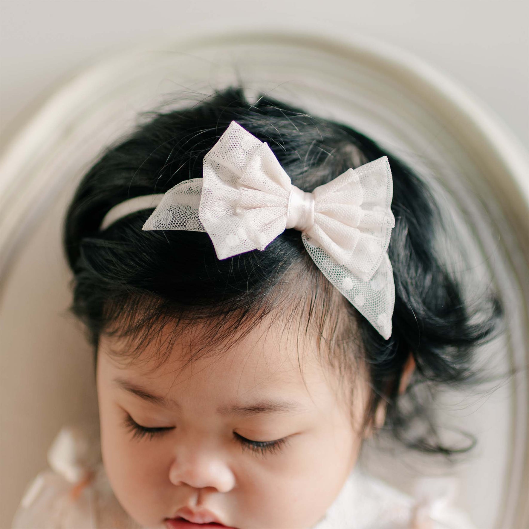 Close-up of the Sophia Lace Bow Headband, featuring delicate ivory lace with embroidered polka dots and a satin center bow, adorning a baby's dark hair.